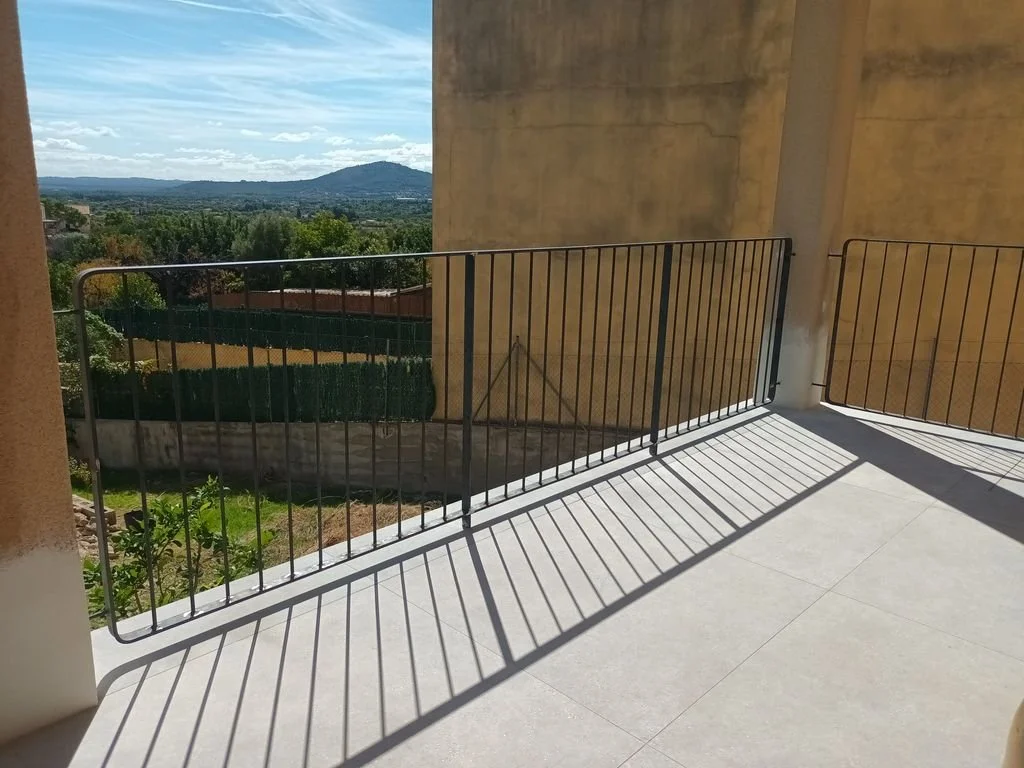 Balcón con barandal de metal y sombra de las barrotes en el suelo, vista de paisaje con árboles, colinas y cielo con nubes.