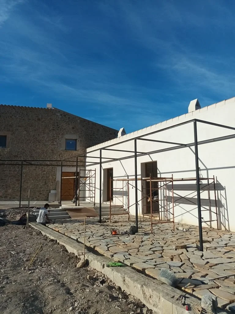 Construcción en progreso en una terraza de piedra en un edificio con paredes blancas y estructura metálica para la cubierta, con herramientas y materiales de construcción.