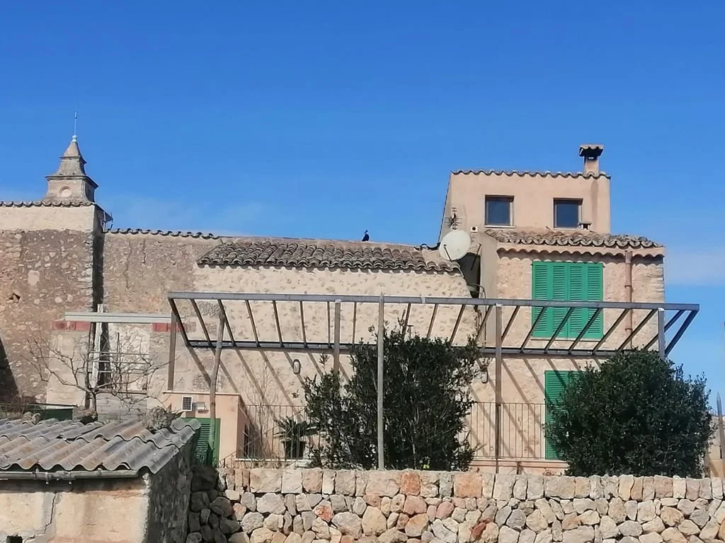 Edificio antiguo de piedra con ventanas de persianas verdes, en un entorno con muro de piedra y estructura metálica en primer plano, cielo despejado.