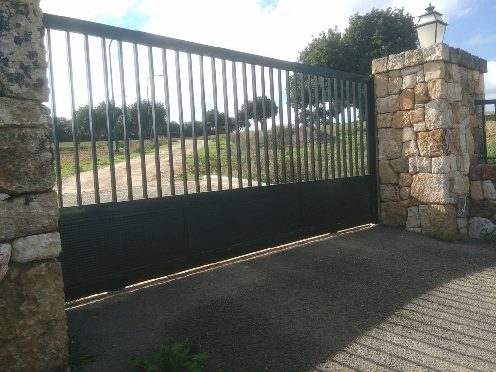 Puerta de reja negra con pilares de piedra y lámpara antigua en la parte superior, en un entorno rural.