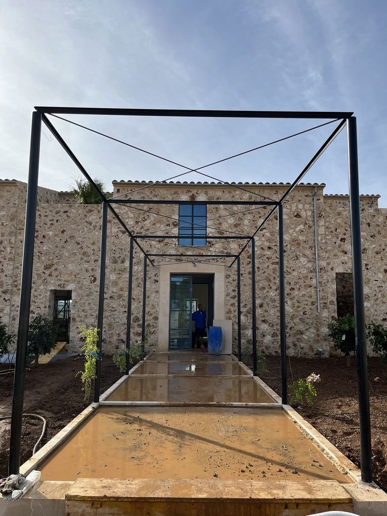 Estructura metálica en construcción frente a una casa de piedra, con un camino de concreto reflectante y un jardin a los lados.