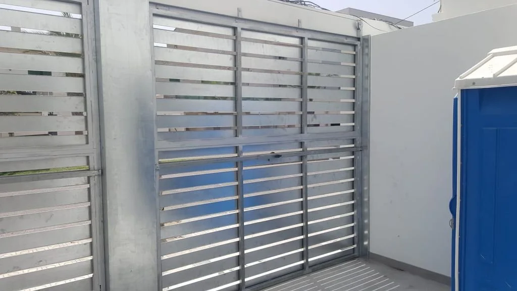 Puerta de reja metálica en un muro blanco, junto a una caseta azul de almacenamiento con techo de acrílico.