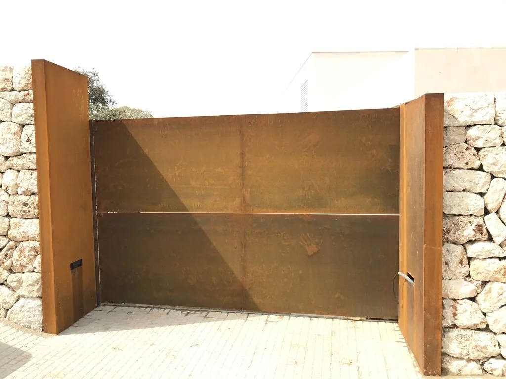 Puerta de entrada de metal con paredes de piedra a cada lado, en un entorno exterior.