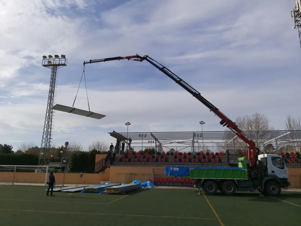 Camión con grúa levantando una estructura o panel en un campo de fútbol.