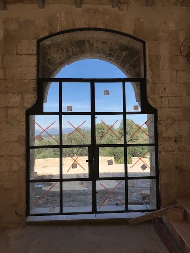 Ventana con marco negro en una pared de piedra, vista a un paisaje con árboles y cielo azul.