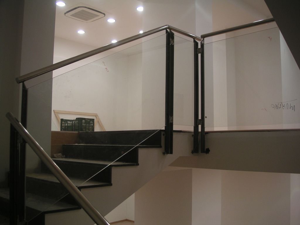 Escalera de interior en construcción con barandales de metal y cristal, paredes blancas y luces en el techo.