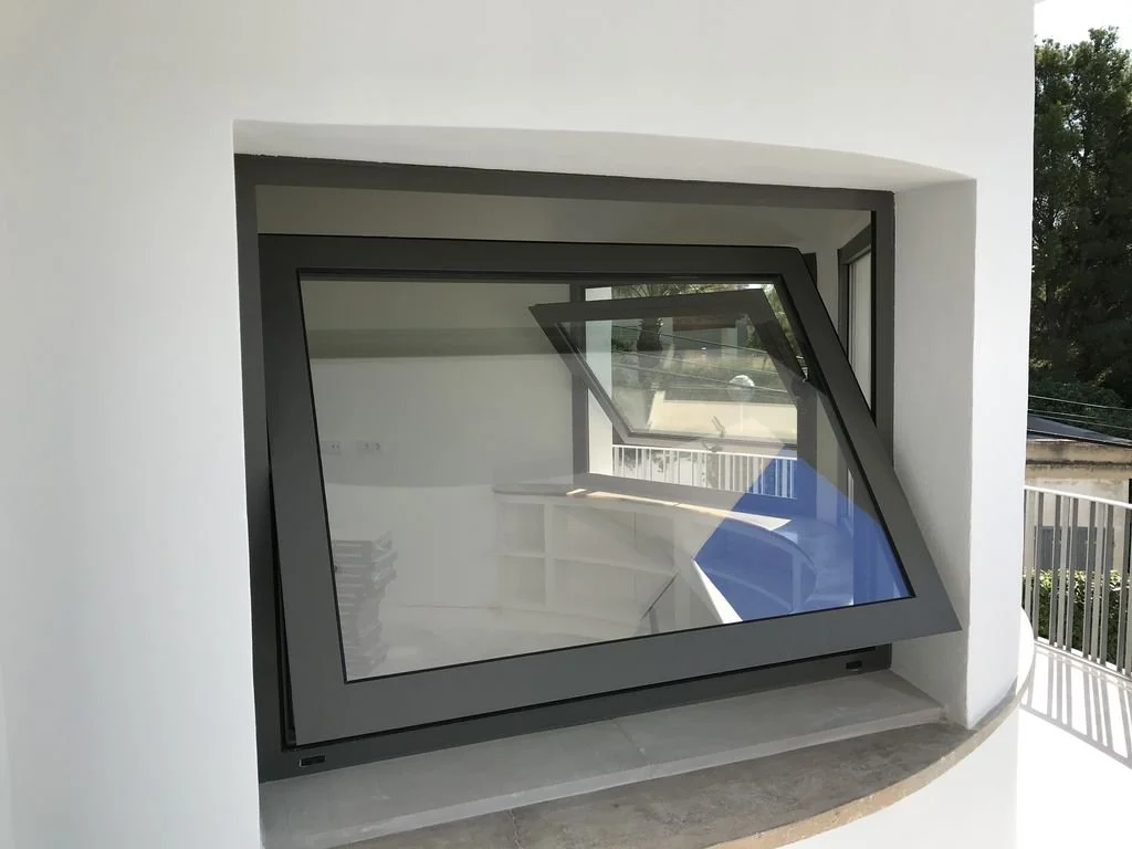 Ventana moderna de diseño inclinada instalada en una pared blanca, con vista a un balcón con barandilla y árboles en el fondo.