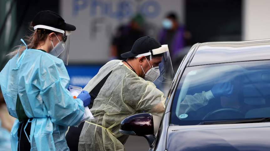 Healthcare workers wearing PPE and masks conduct a COVID-19 test on a person inside a vehicle at a drive-thru testing site.
