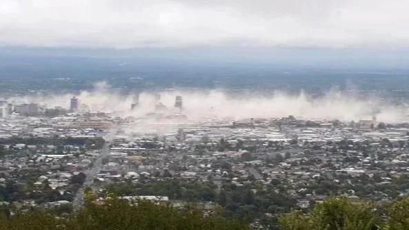 Christchurch during the Earthquake view under cloudy sky with smoke or fog billowing from buildings in the distance.