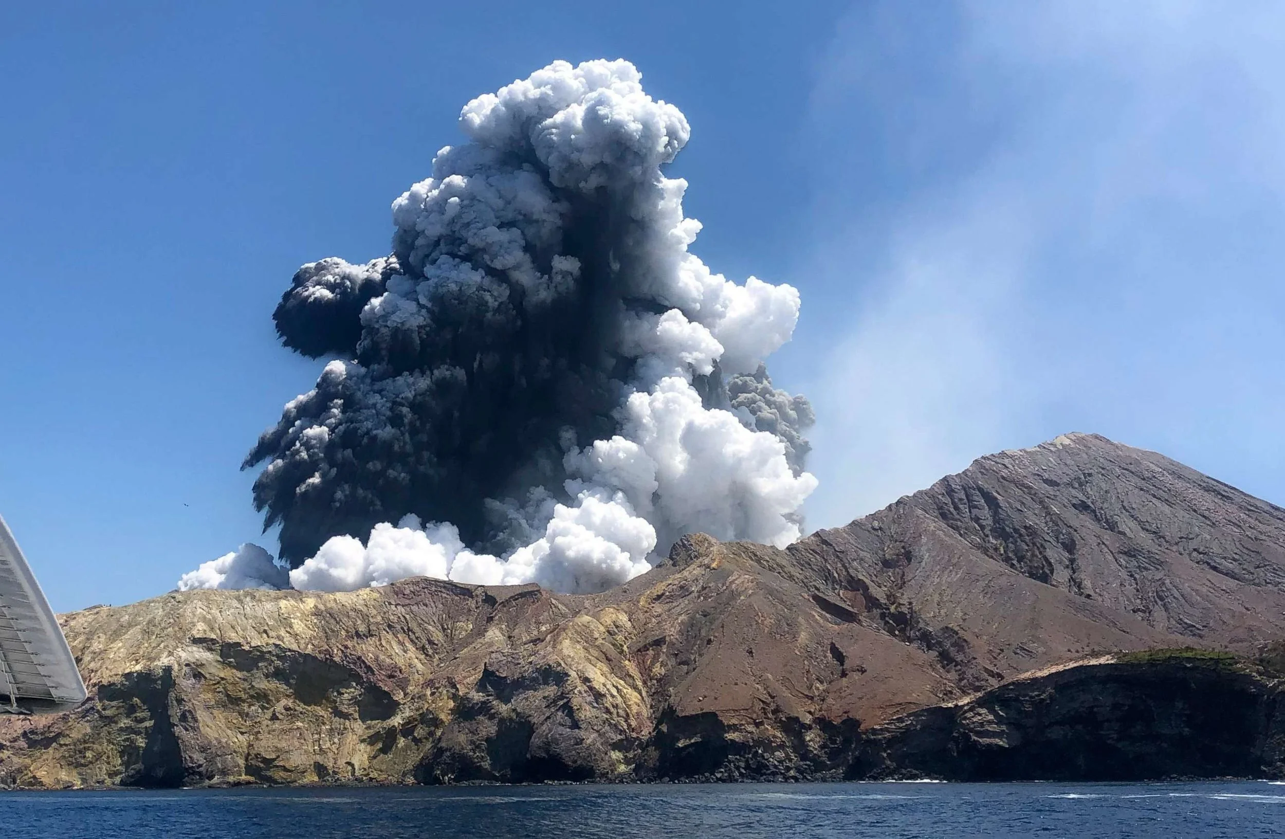 A White Island eruption spewing ash and smoke into the sky from a mountain near a body of water.