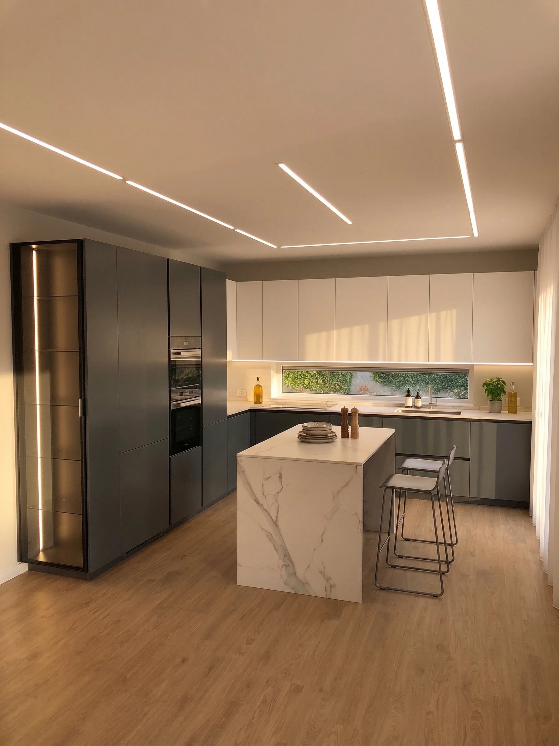 Cucina moderna con armadi blu e bianco, isola centrale in marmo bianco con venature grigie, due sgabelli in metallo e sedile in pelle, grande finestra che mostra un giardino verde.