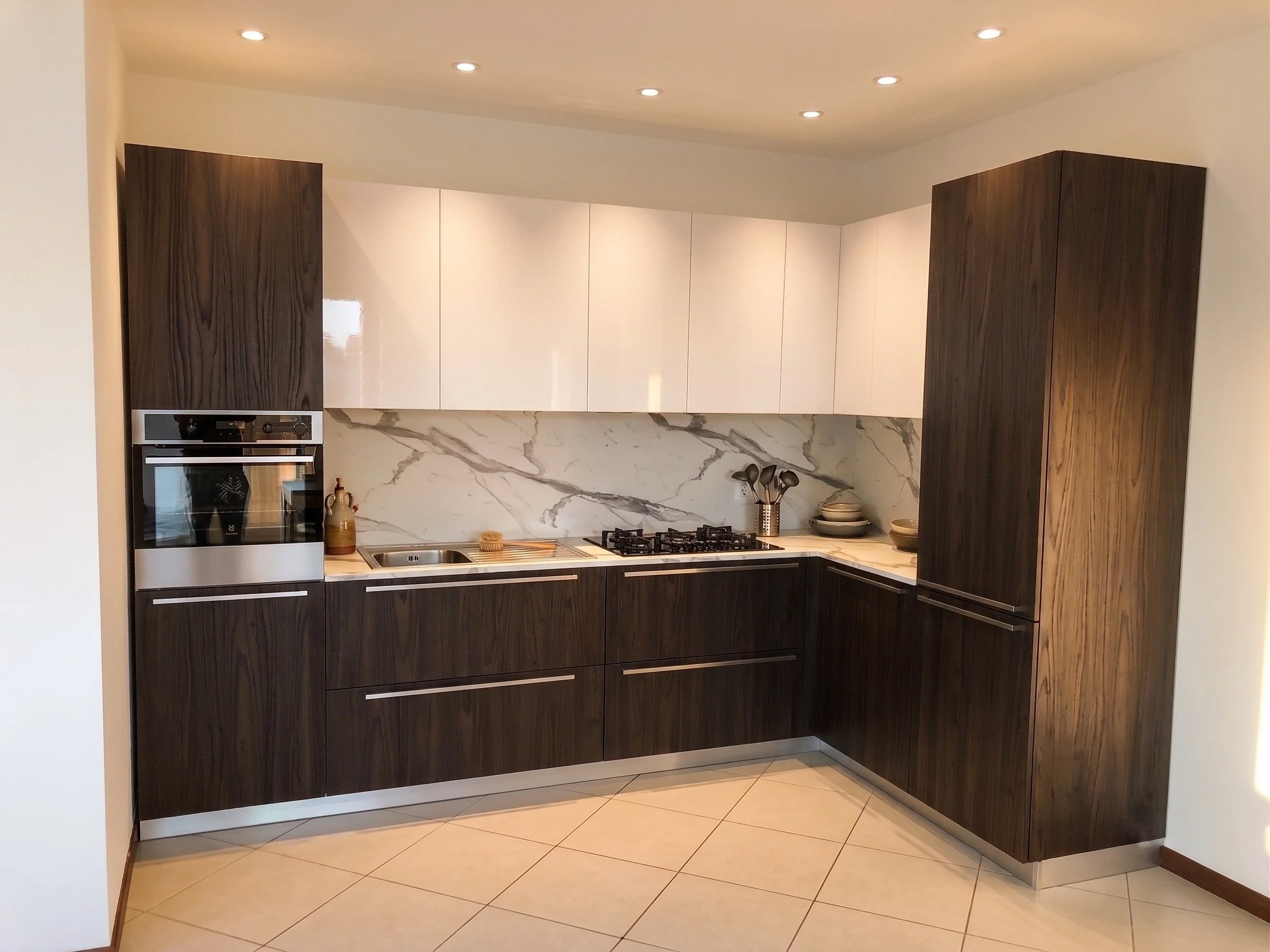 Cucina moderna con armadi in legno scuro, parete in marmo bianco con venature grigie, piano cottura a gas, forno integrato, e alcuni utensili e stoviglie.