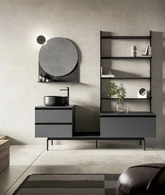 Mobili da bagno moderni con lavabo rotondo, specchio rotondo, scaffali neri, vaso con pianta in vetro e rivestimento grigio.