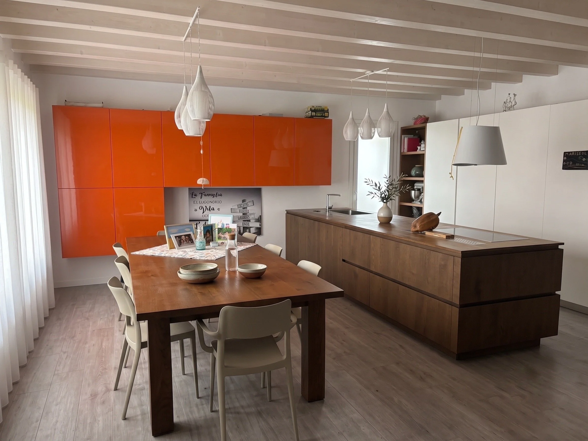 Interno di una cucina moderna con pareti bianche, controsoffitto con travi di legno chiaro, un tavolo in legno allungabile con sedie bianche, arredamento con armadi in legno scuro e arancione lucido, illuminazione sospesa con lampade a goccia bianche, piante e foto sulla parete, credenza in legno.