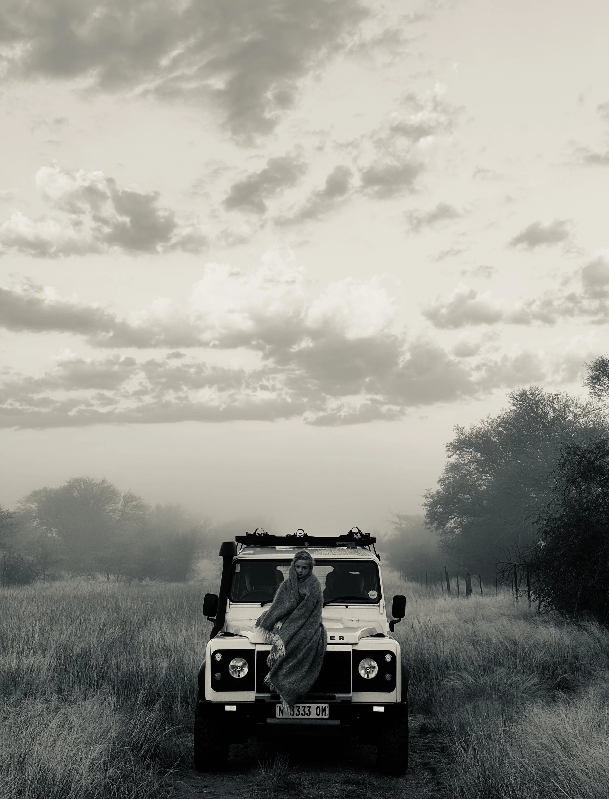 A young boy, wrapped in a soft mohair blanket, sitting on the hood of a white Land Rover Defender on a grassy farm track, with trees and cloudy sky in the background, black and white photo.