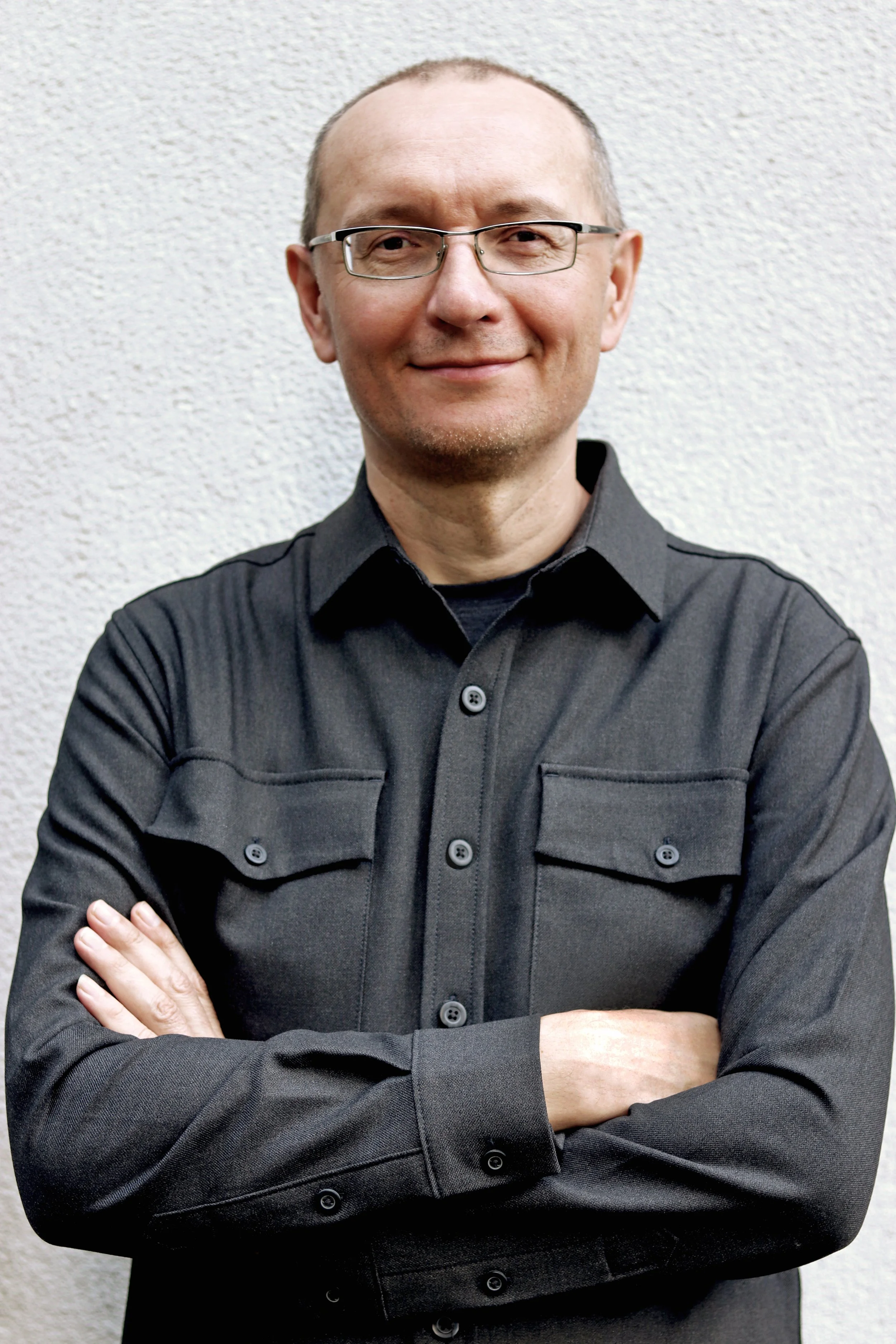 A man with short hair, glasses, and a light beard wearing a black button-up shirt with crossed arms standing in front of a textured white wall.