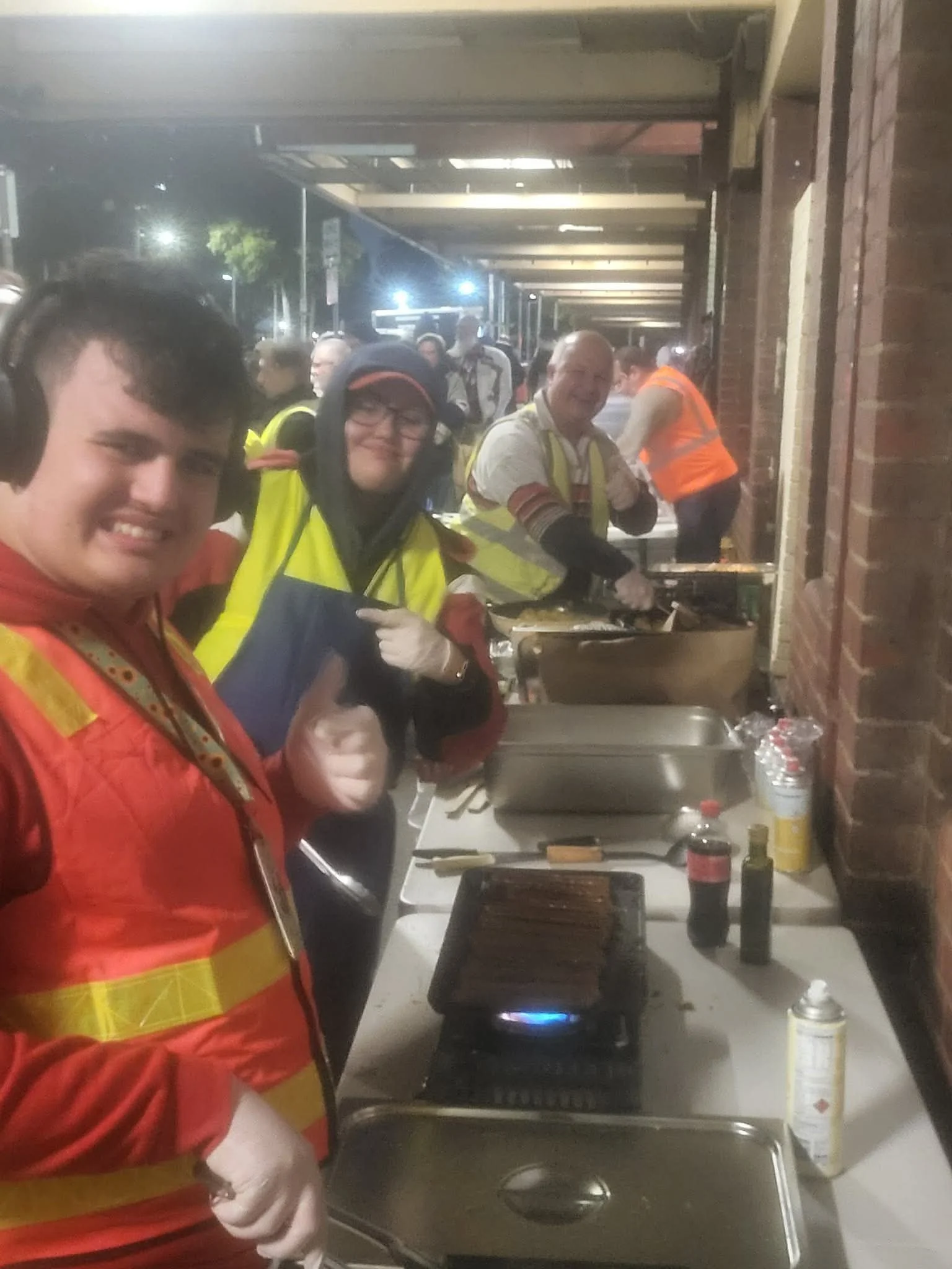 People cooking food on outdoor grills at a night event, wearing reflective vests, with tables, food, and condiments visible.