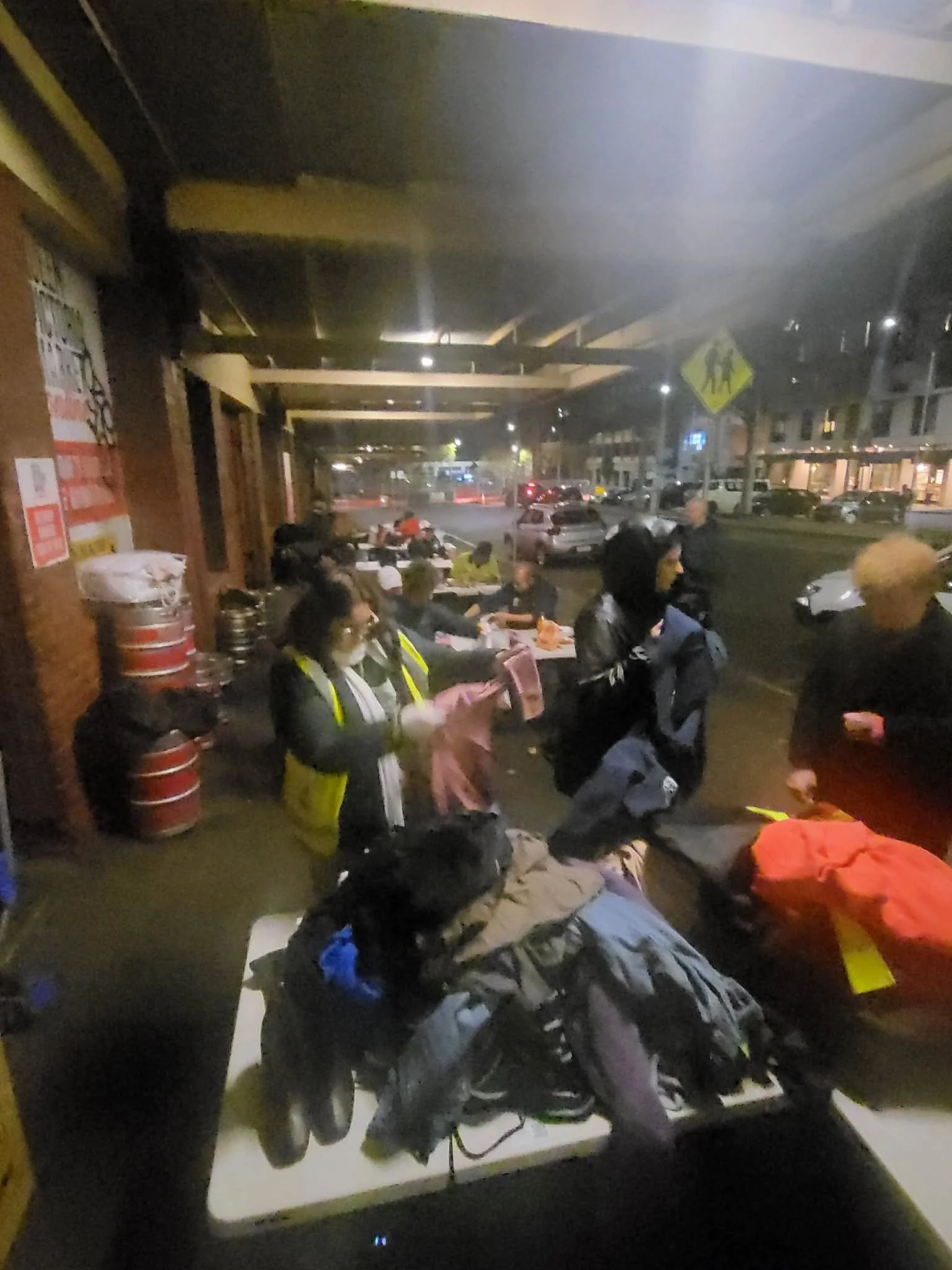 People sitting on a sidewalk at night, some wearing masks, with parked cars and a building in the background.
