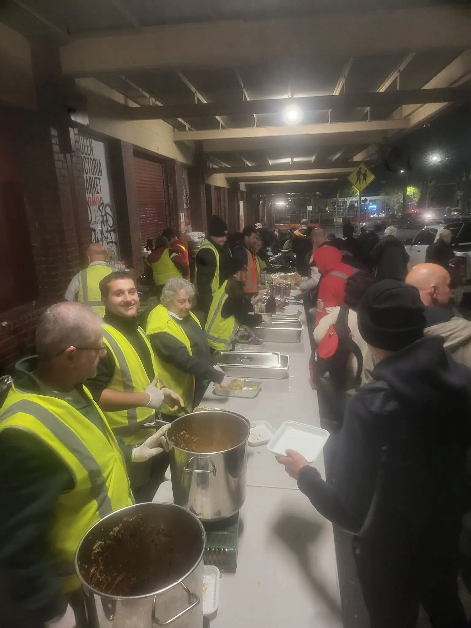 People wearing yellow safety vests preparing food at a community meal event under an overpass at night.