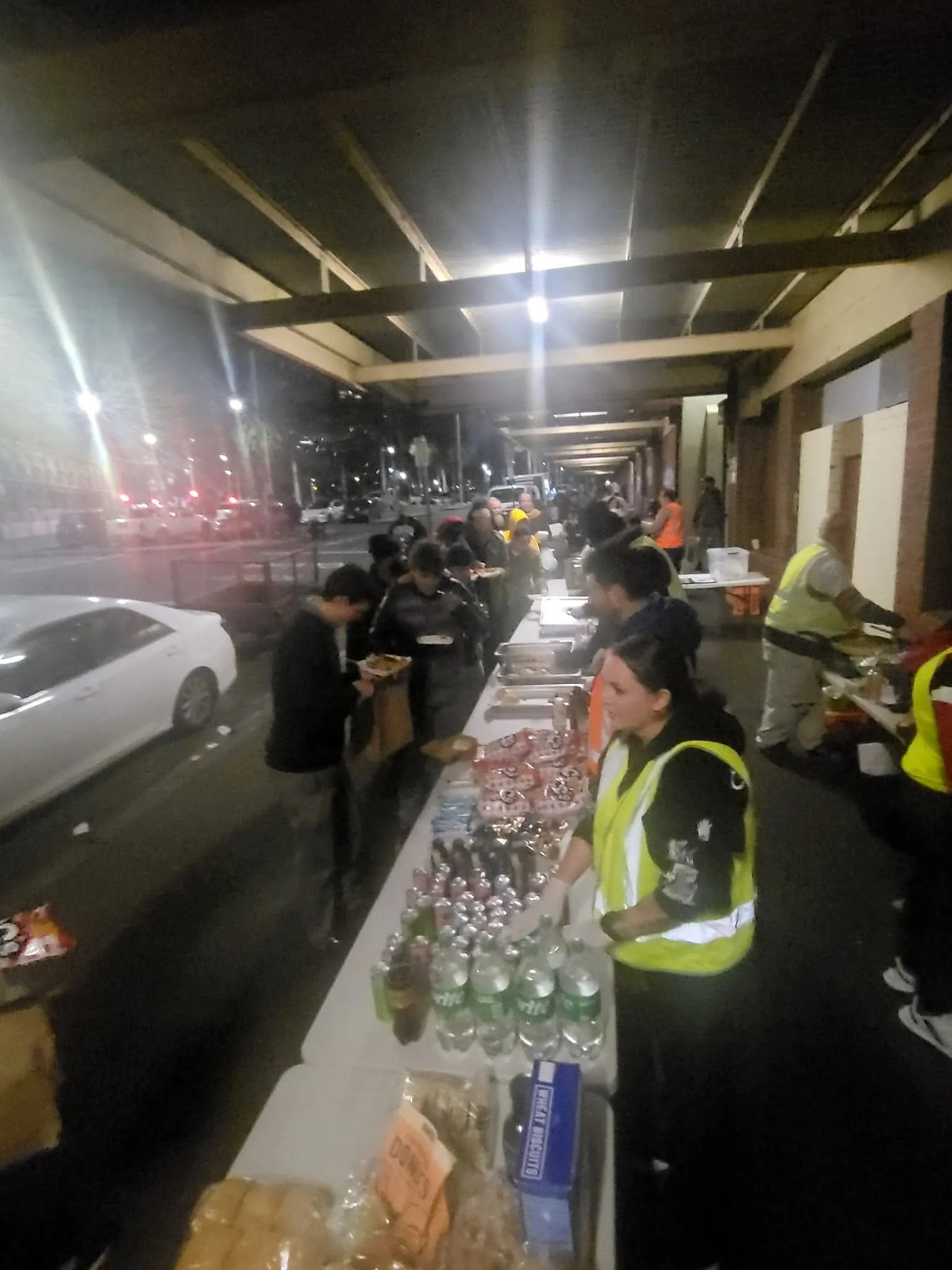 People standing in line at a table with various food and drinks outdoors at night.