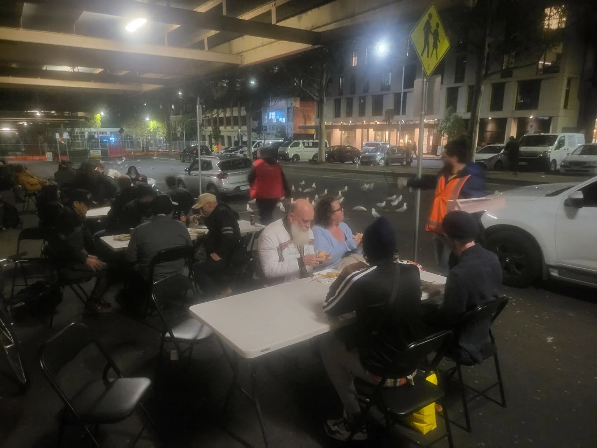 People seated at tables outside a restaurant at night, with a parking lot and buildings in the background, and a traffic sign indicating a pedestrian crossing.