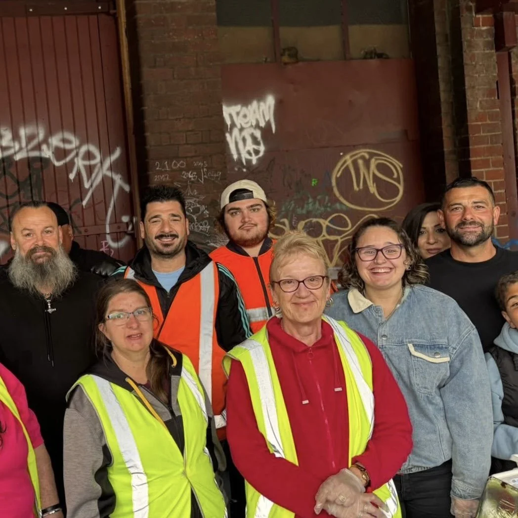 Group of nine diverse people, some wearing high-visibility safety vests, posing together in front of a graffiti-covered brick wall.