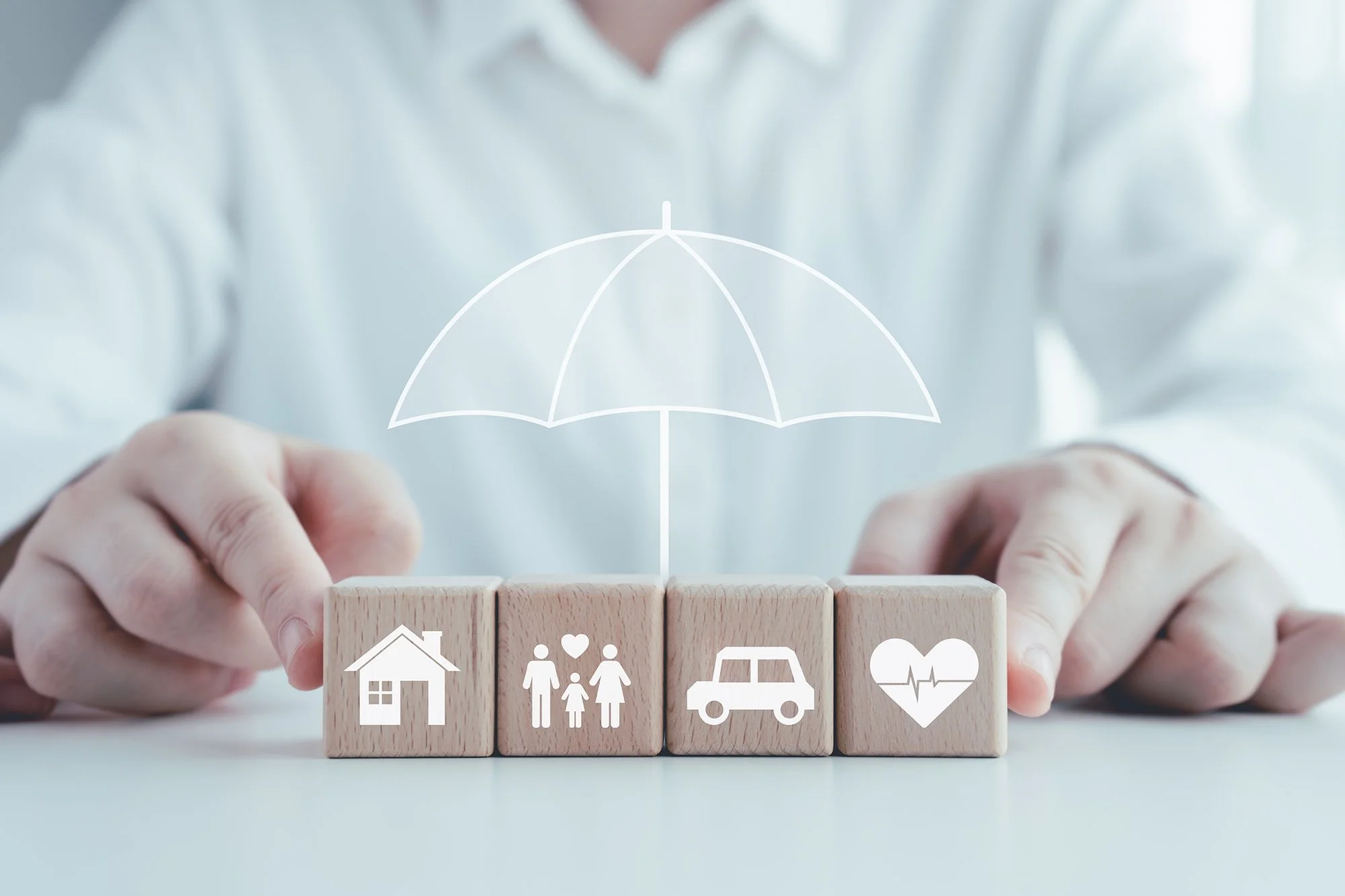 A person in a white shirt arranging wooden blocks with icons representing a house, family, car, and heart health, with a digital umbrella symbol above.
