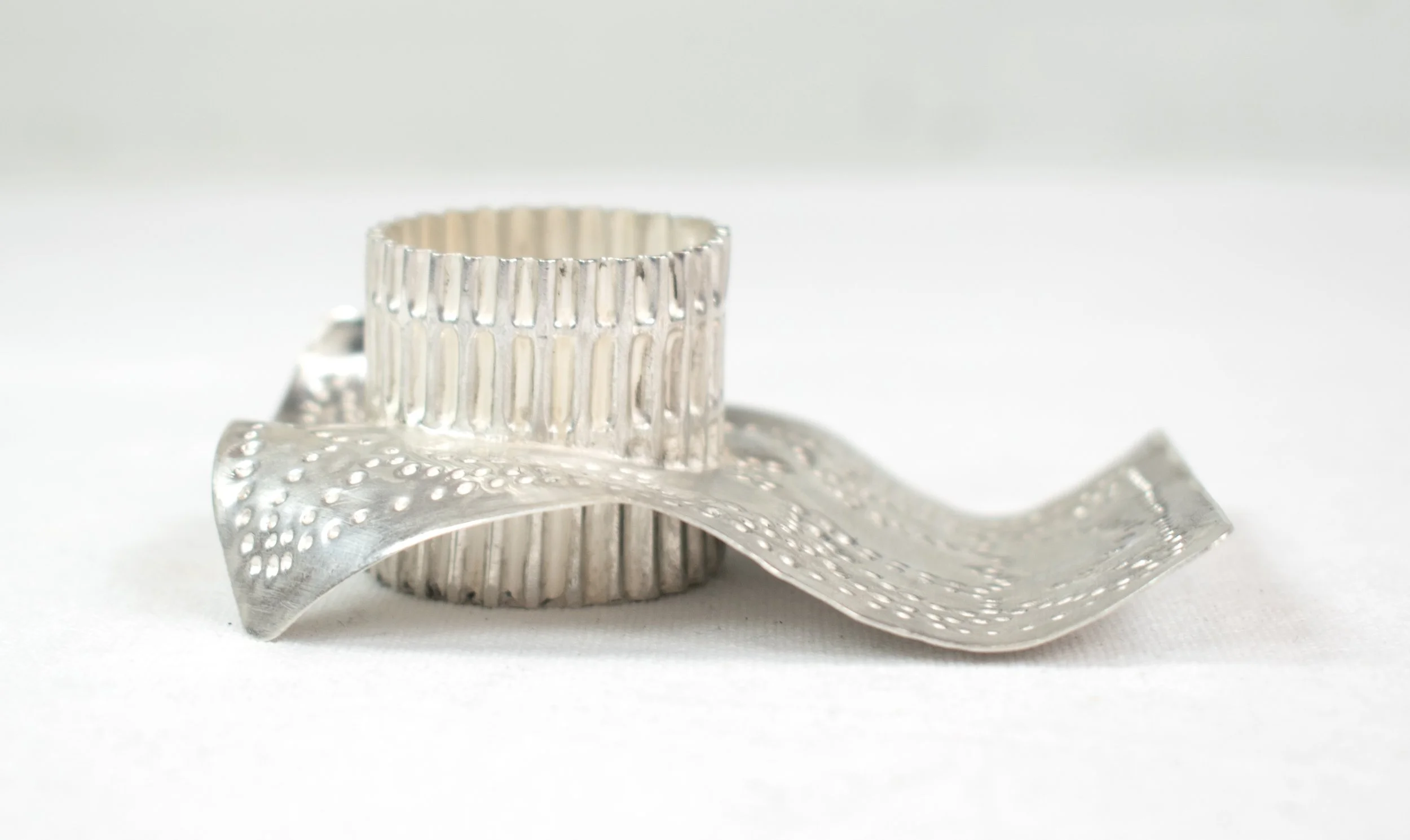 A silver ring with vertical ridges, placed on top of a silver ribbon with dotted patterns on a white background.