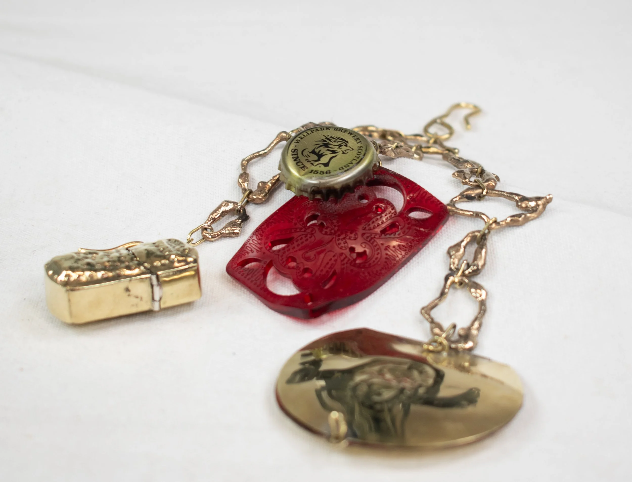 Close-up of a necklace with a bottle cap pendant featuring a lion and the text 'WELLPARK BREWERY SCOTLAND 1556' on a white surface.