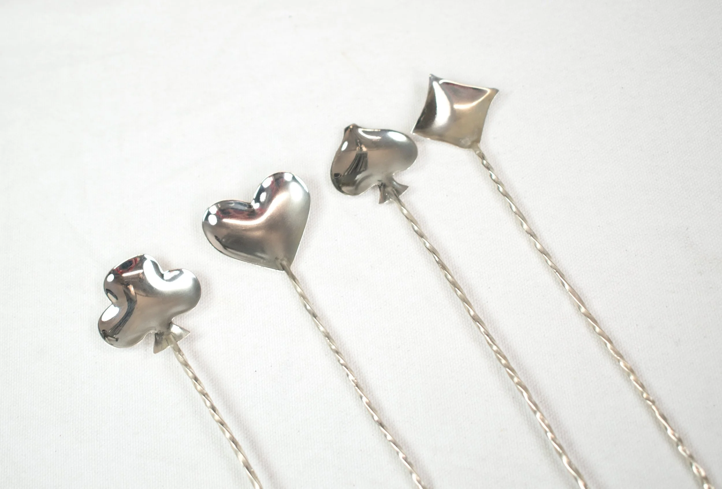 Four metallic drink stirrers with decorative tops in the shape of a heart, star, cube, and flower, laid out on a white surface.