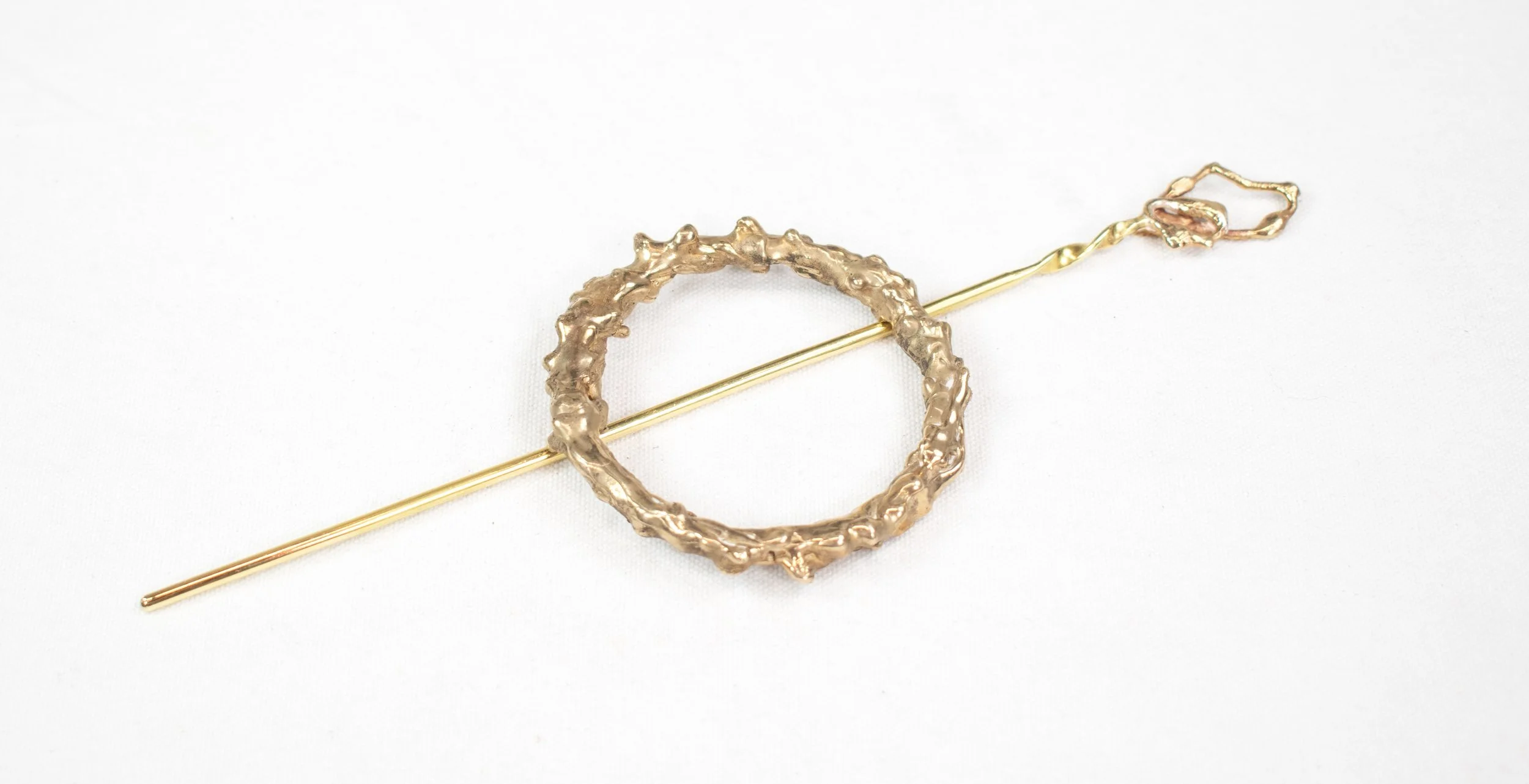 Gold jewelry piece with a textured circular element and a long, straight pin on a white background.