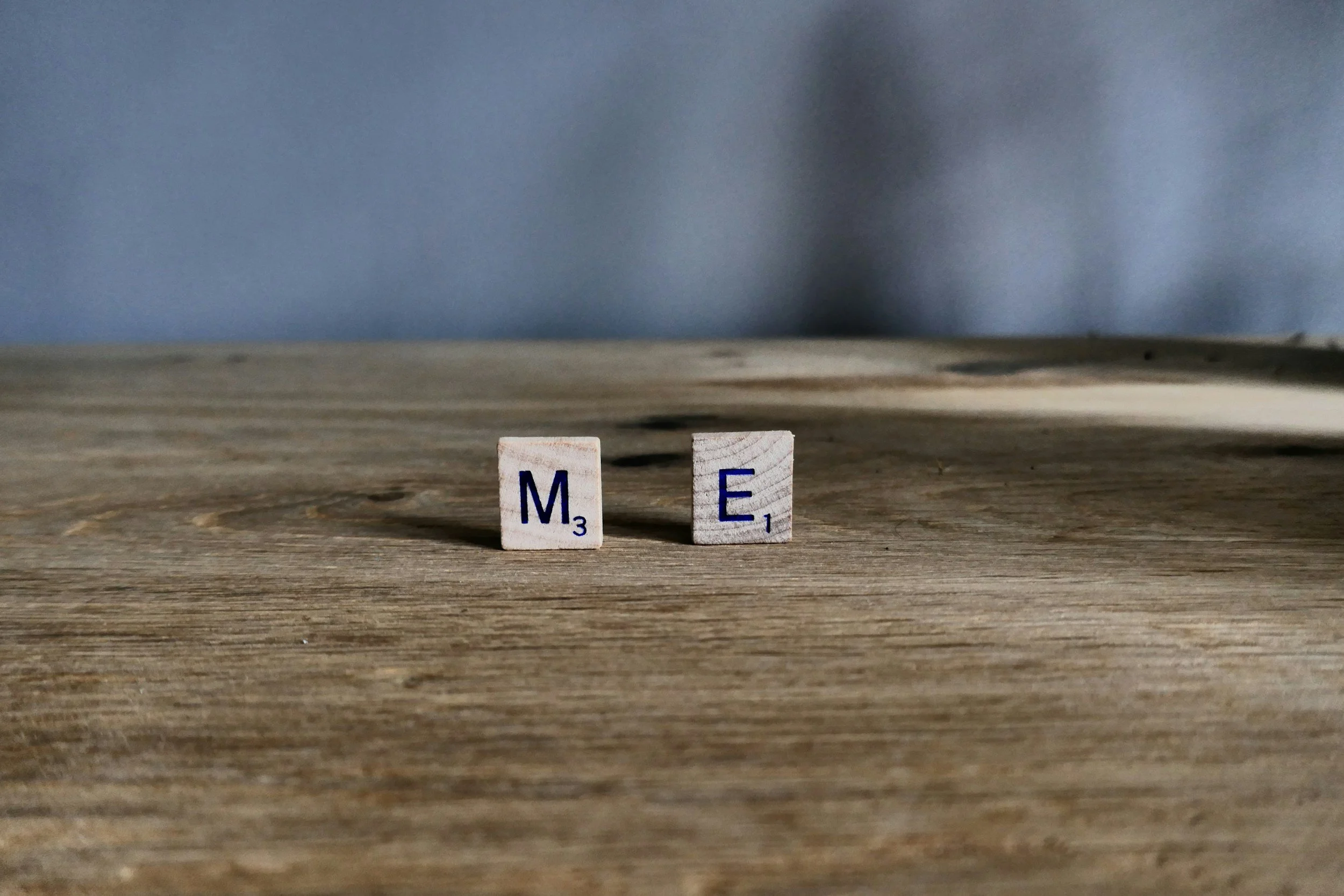 Close-up of two wooden Scrabble tiles on a wooden surface spelling out 'ME' with a blue-gray background.