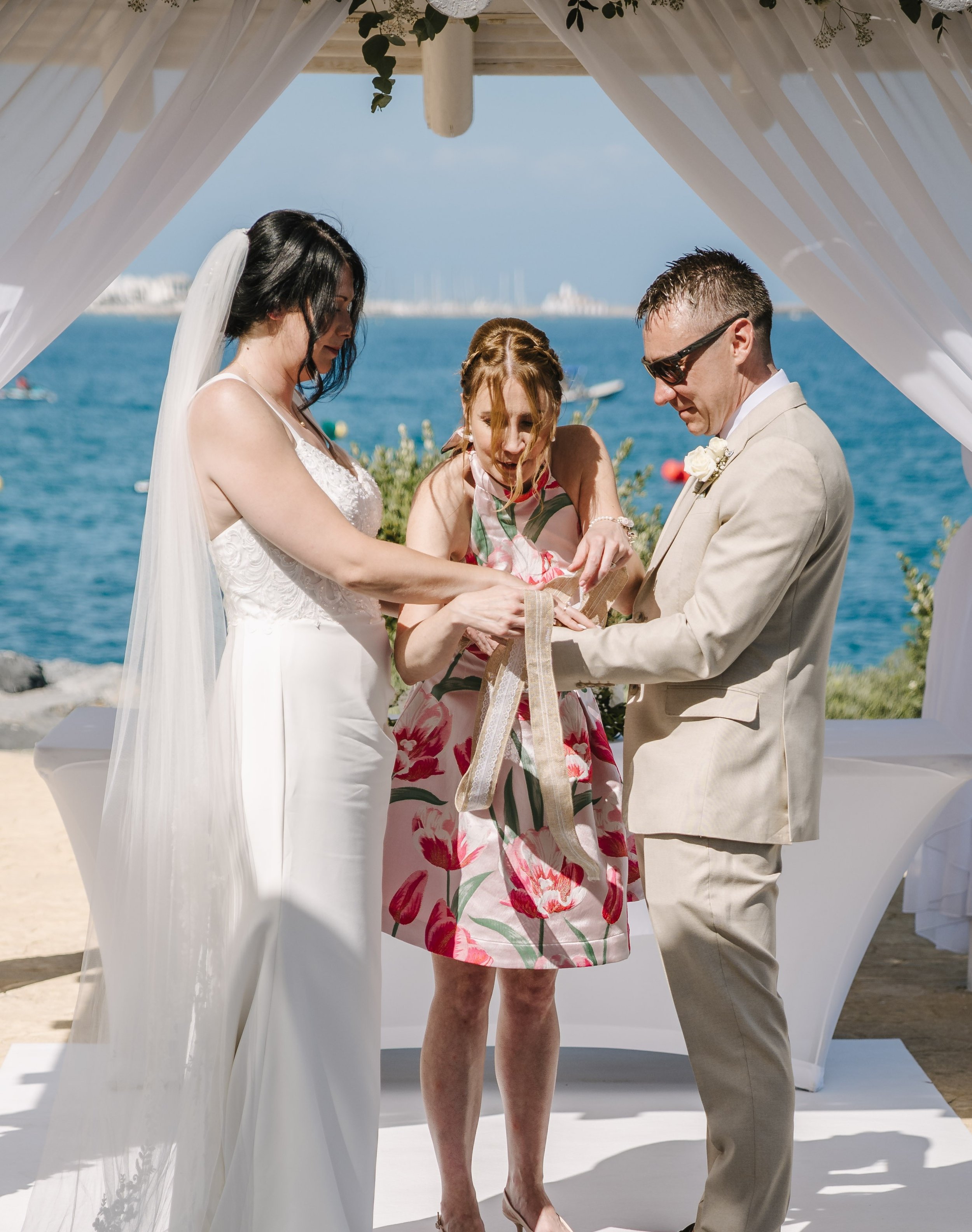 A wedding ceremony outdoors by the water with three people standing under a white canopy. The woman in the middle is in a floral dress, the woman on the left is in a white wedding gown with a veil, and the man on the right is in a beige suit with sunglasses.