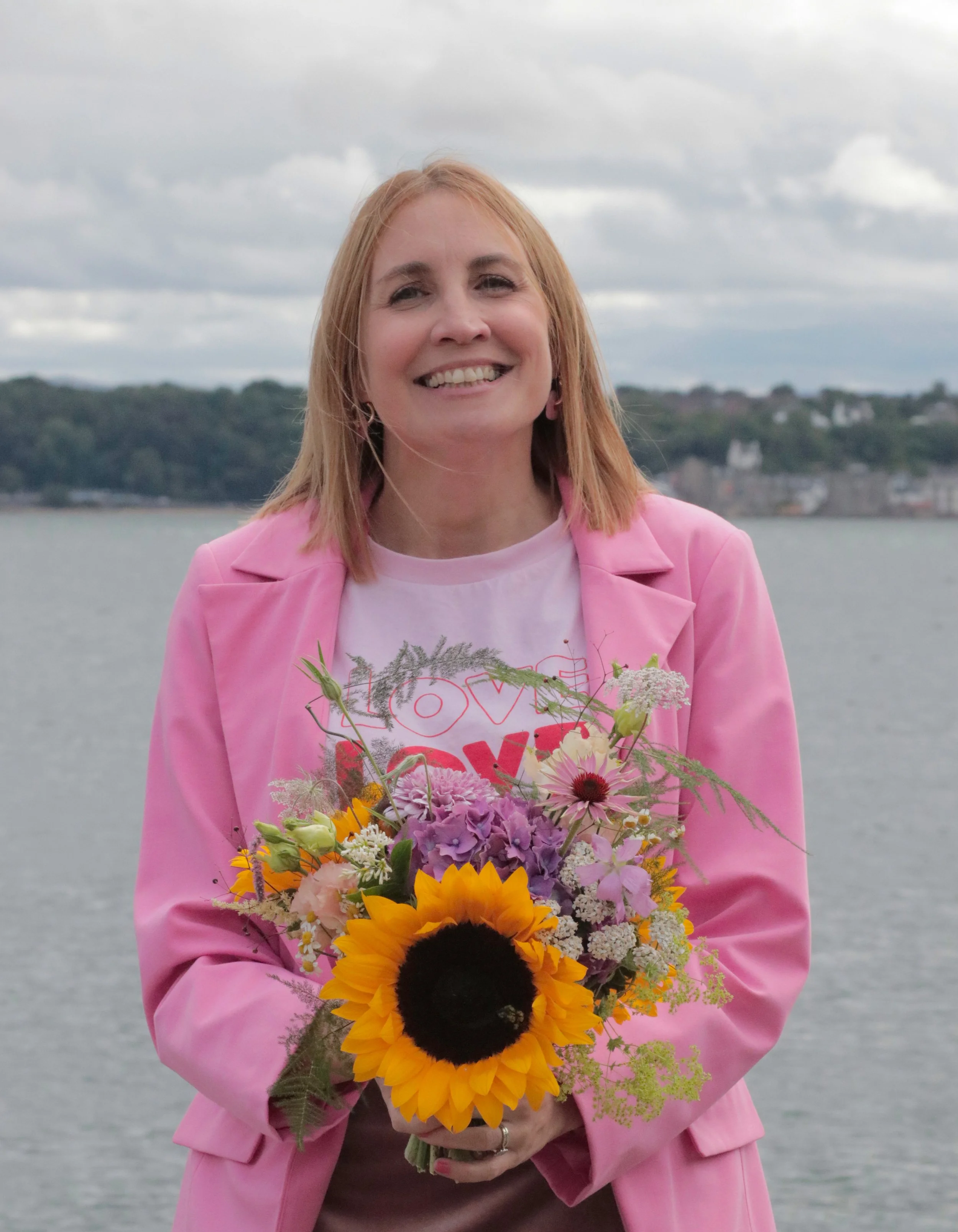 Debbie Anne, a woman with shoulder-length blonde hair and a pink blazer holding a colourful bouquet of flowers in front of a body of water and cloudy sky.
