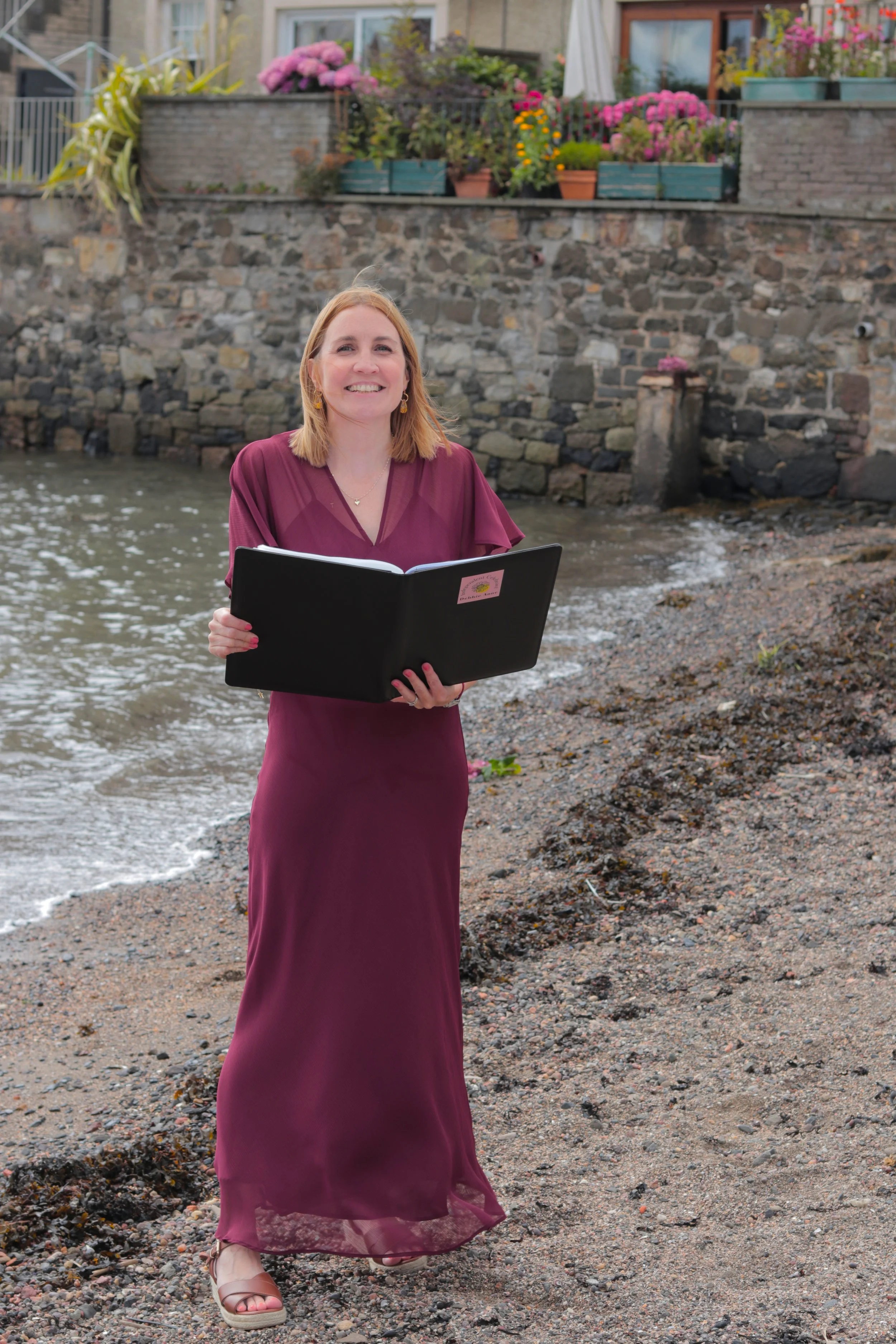 A woman in a burgundy dress standing on a rocky beach holding a black folder, with a stone wall and colorful flowers in planters in the background.
