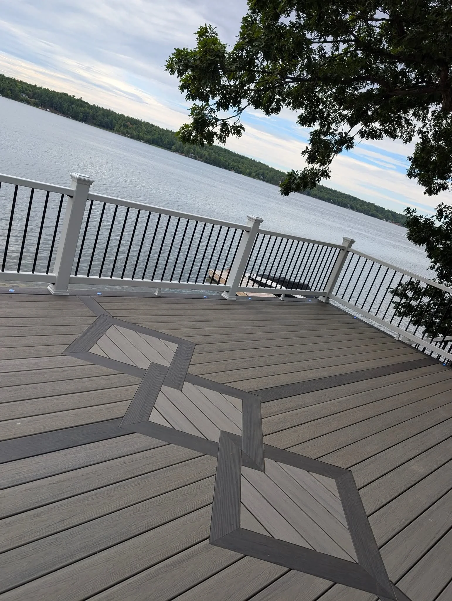 View of a lake seen from a wooden deck with a metal railing, trees overhanging, and a cloudy sky.