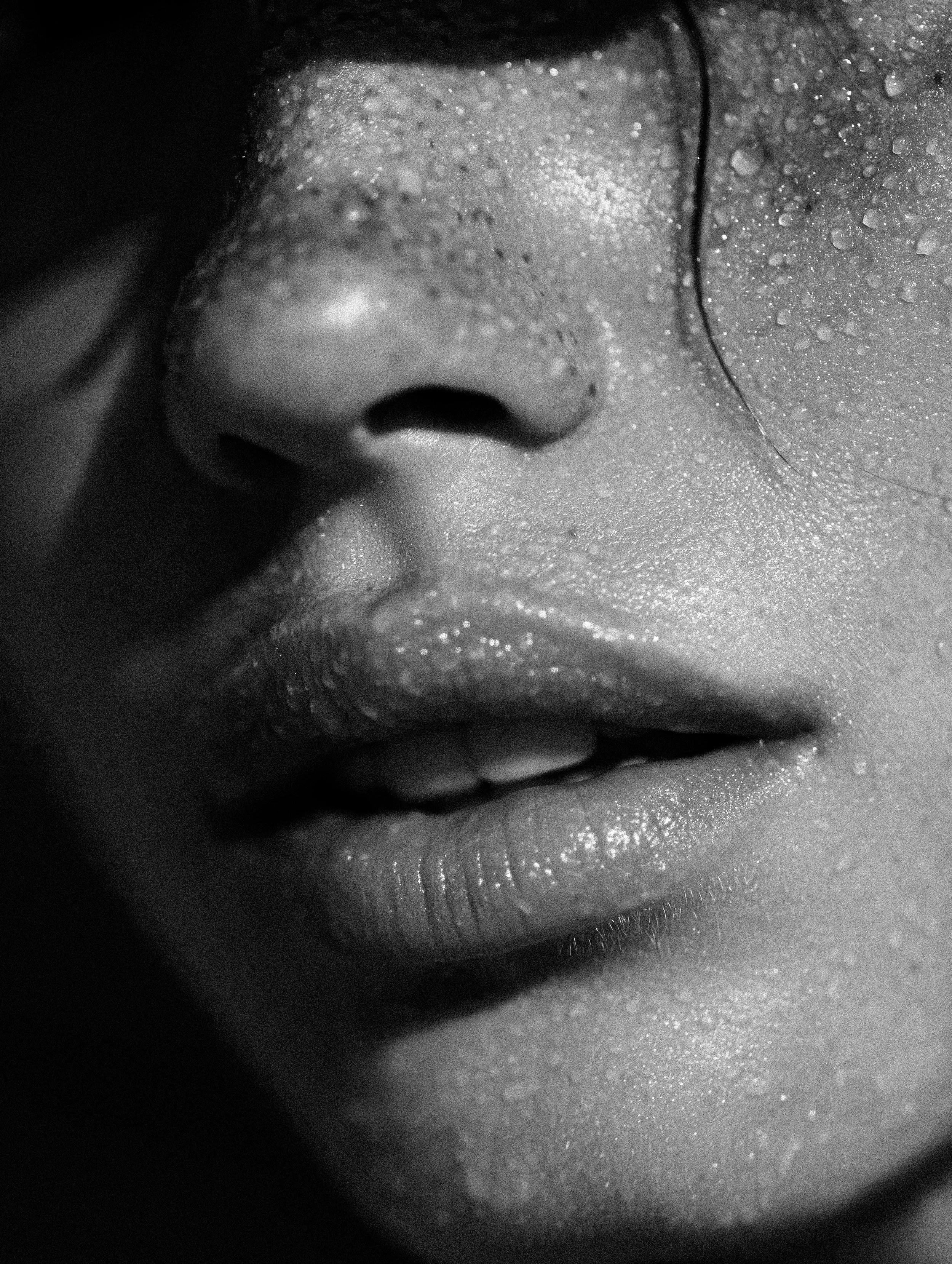 Close-up black-and-white photo of a person's face, focusing on the closed eye, nose, and lips, with water droplets on the skin.