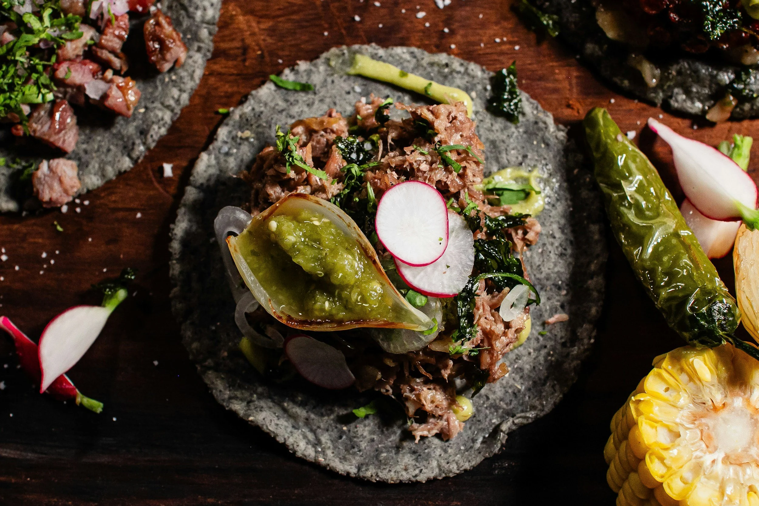 Close-up of a Mexican dish, featuring a stone serving platter with finely chopped cooked meat garnished with herbs, radish slices, and a grilled green pepper.
