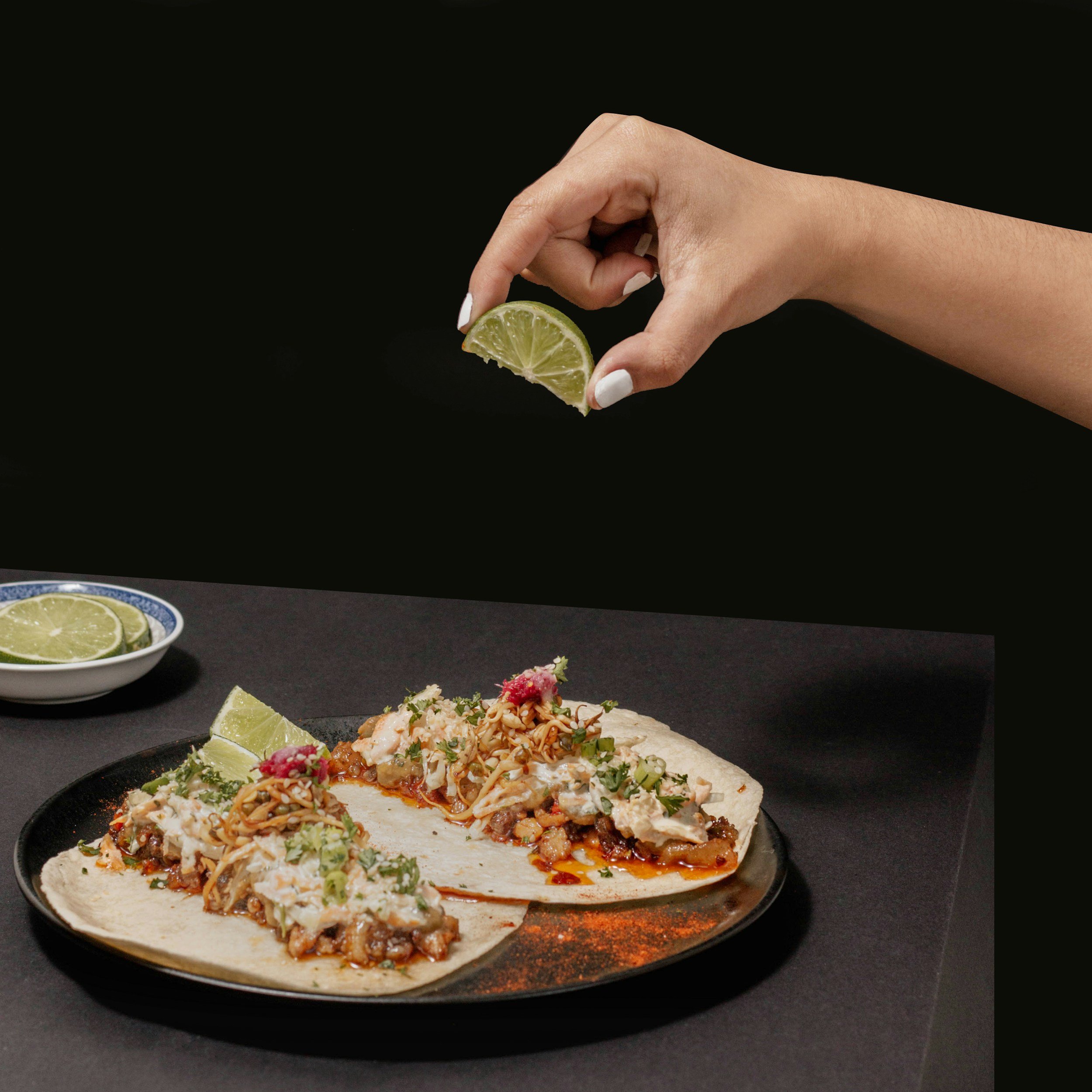 A hand holding a lime wedge over a plate of tacos filled with meat, vegetables, and garnishes, with additional lime wedges on a small bowl nearby