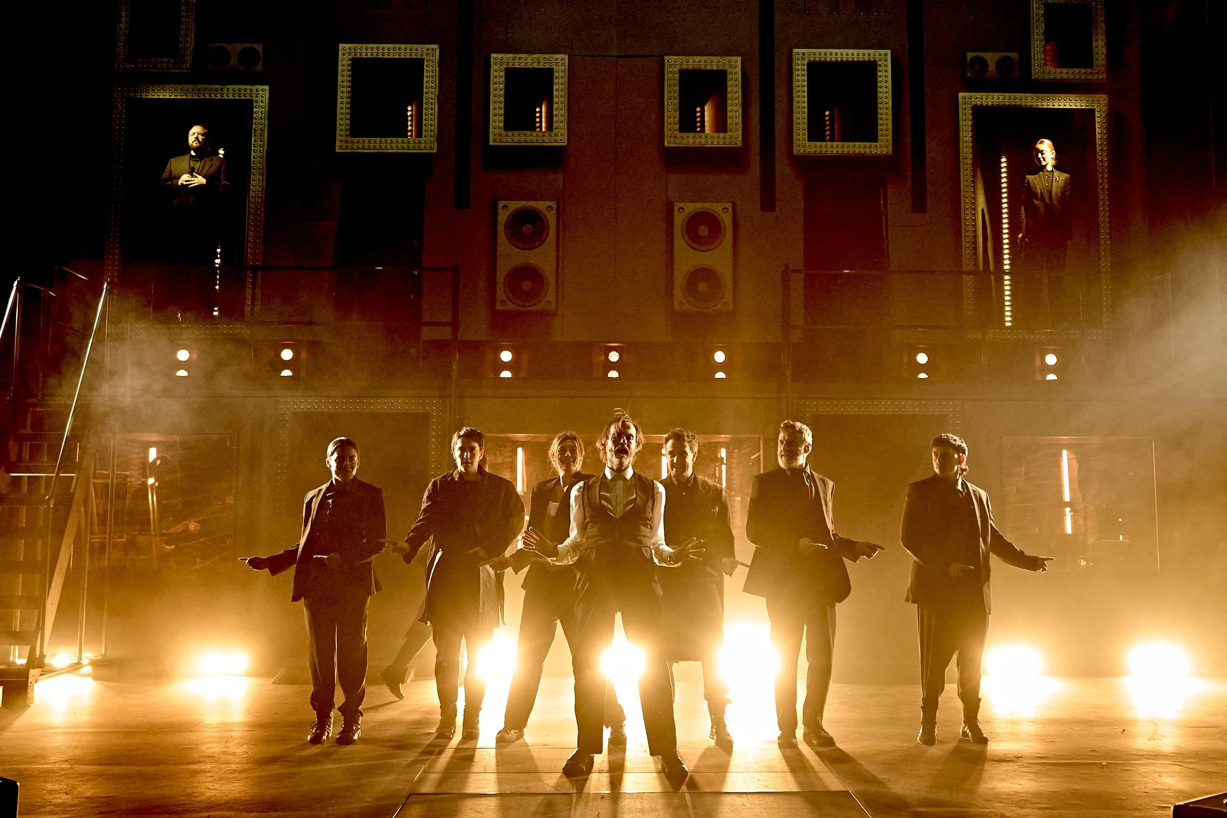 A group of performers dressed in formal tuxedos standing on a stage illuminated by bright warm lights, with a dark background featuring framed mirrors and speakers, creating a dramatic atmosphere.