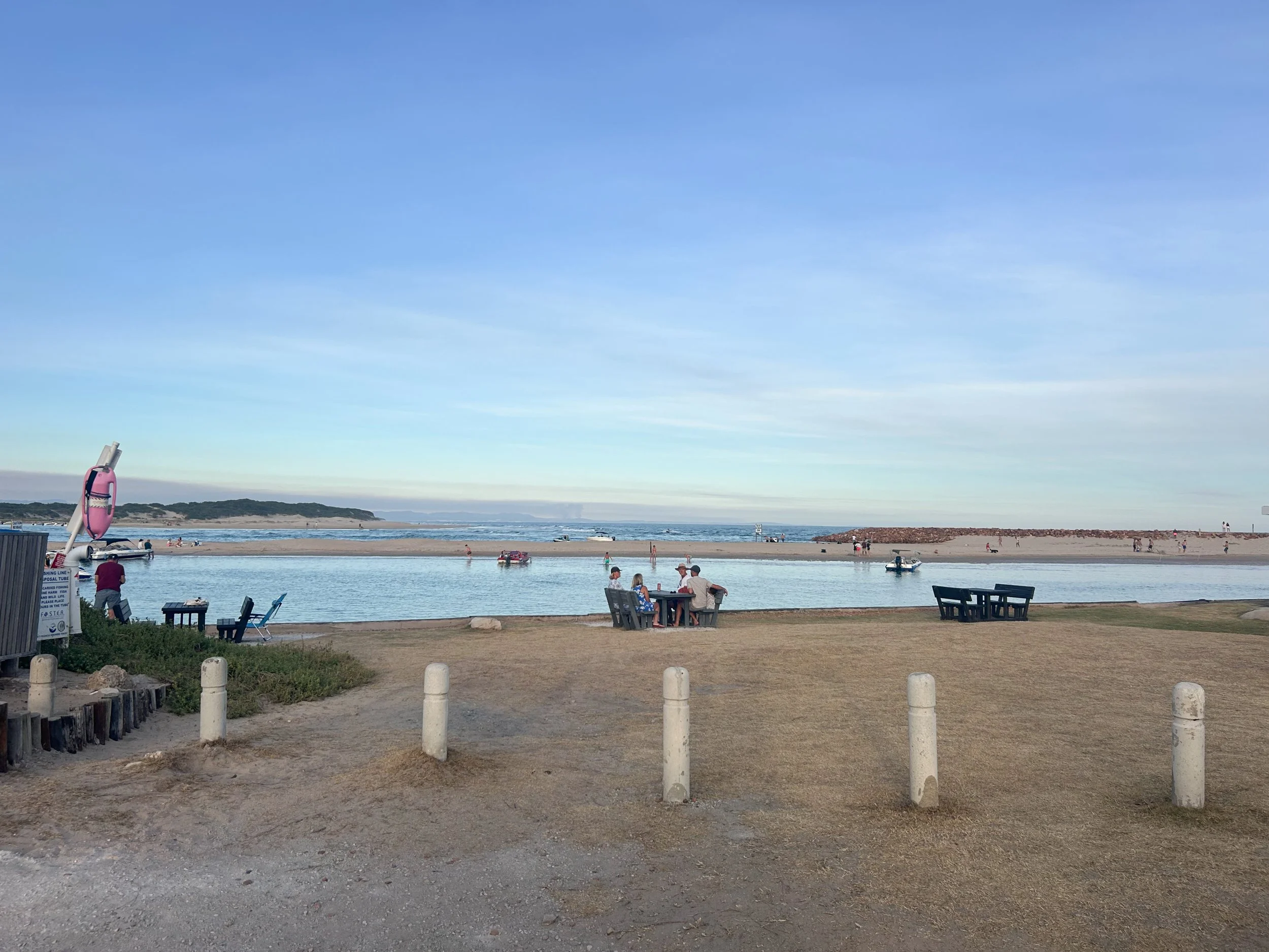 Beach with a pier, kayaks, and people relaxing near the water, under a blue sky with some clouds.