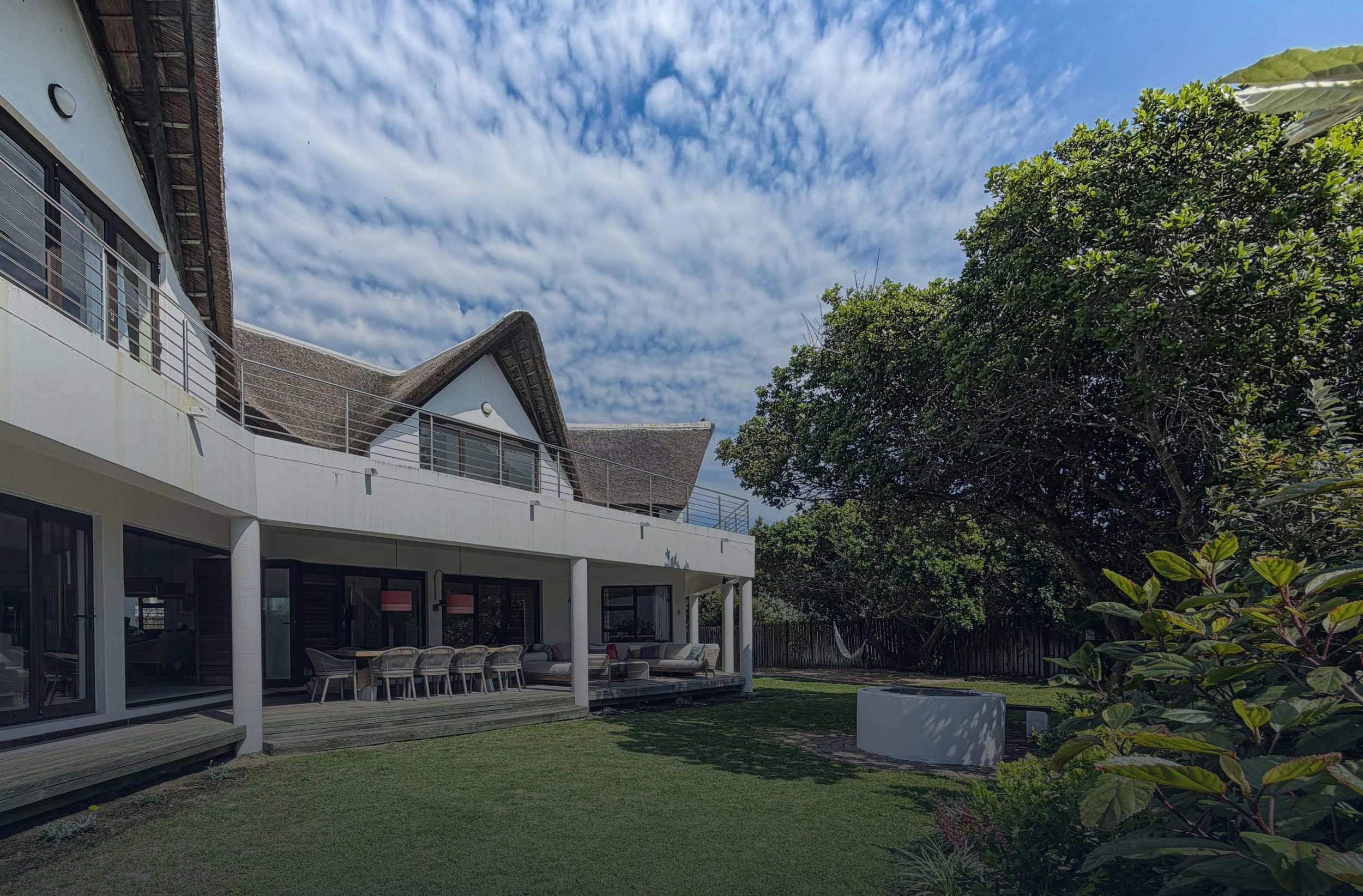 Modern white house with large balcony, outdoor seating area, lush green lawn, surrounded by trees, under a partly cloudy sky.
