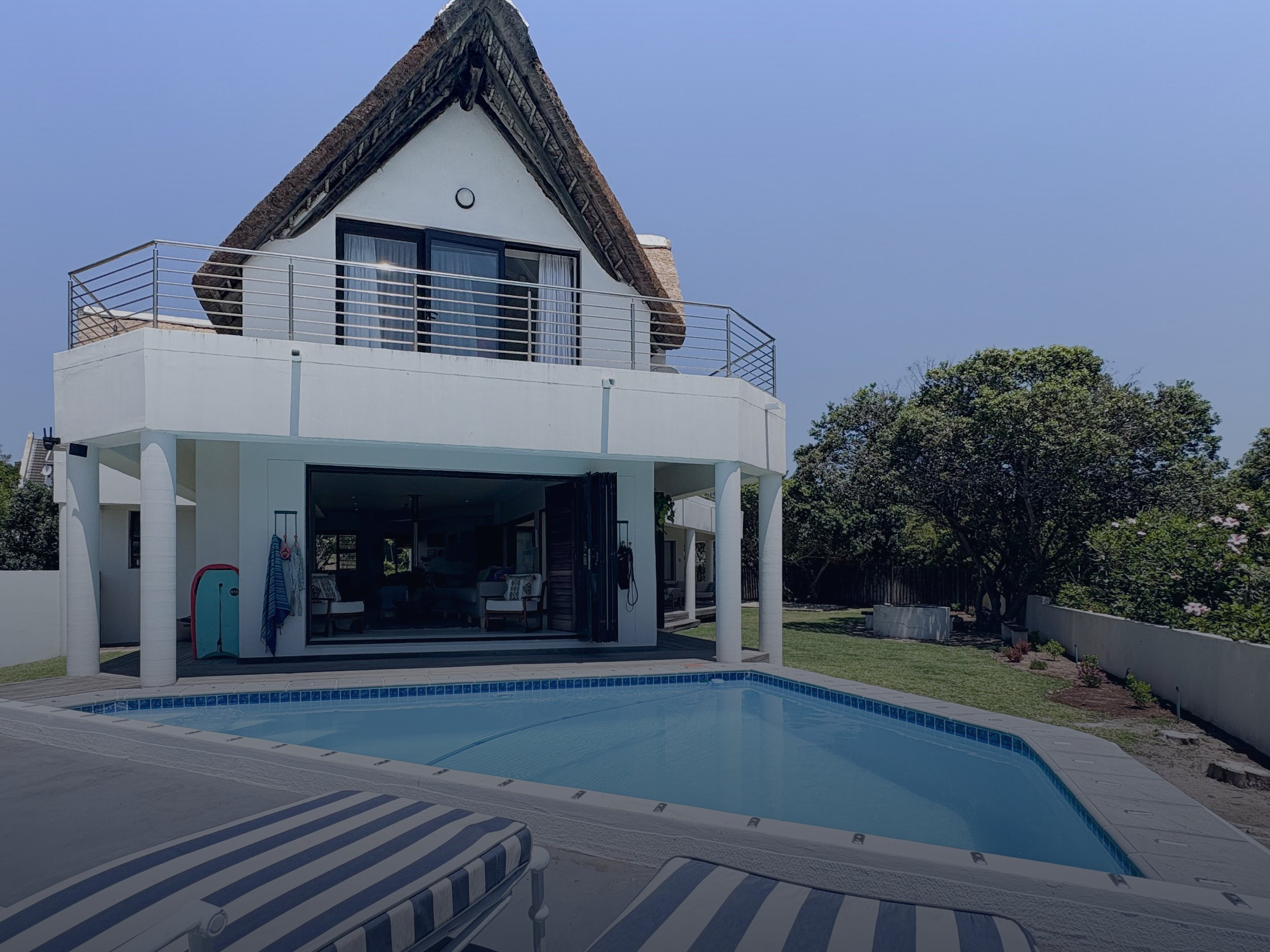 A modern house with a pool in the backyard. The house has a white exterior with a balcony and large glass doors. There are two striped lounge chairs near the pool, and the yard is landscaped with trees and shrubs under a clear blue sky.
