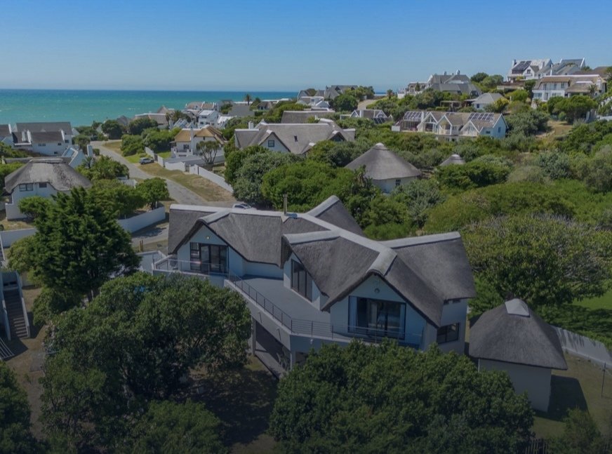 Aerial view of houses with thatched roofs, lush green trees, and a distant view of the ocean under a clear blue sky.
