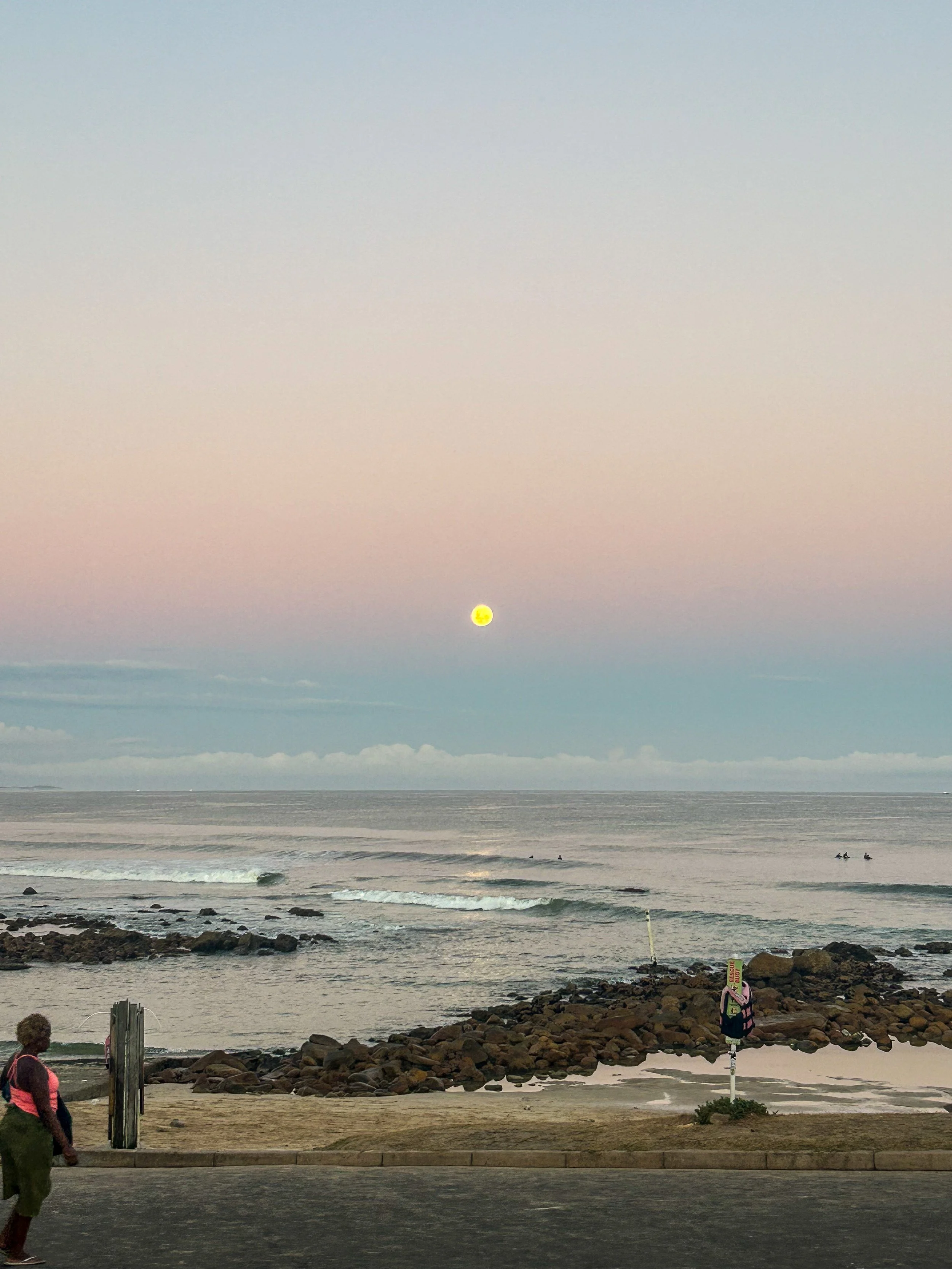 A beach scene at sunset with the sun near the horizon over the ocean. The sky transitions from light to darker shades. Waves are gently crashing onto the shore, and there are rocks along the coastline. Two people are visible on the rocky part of the 