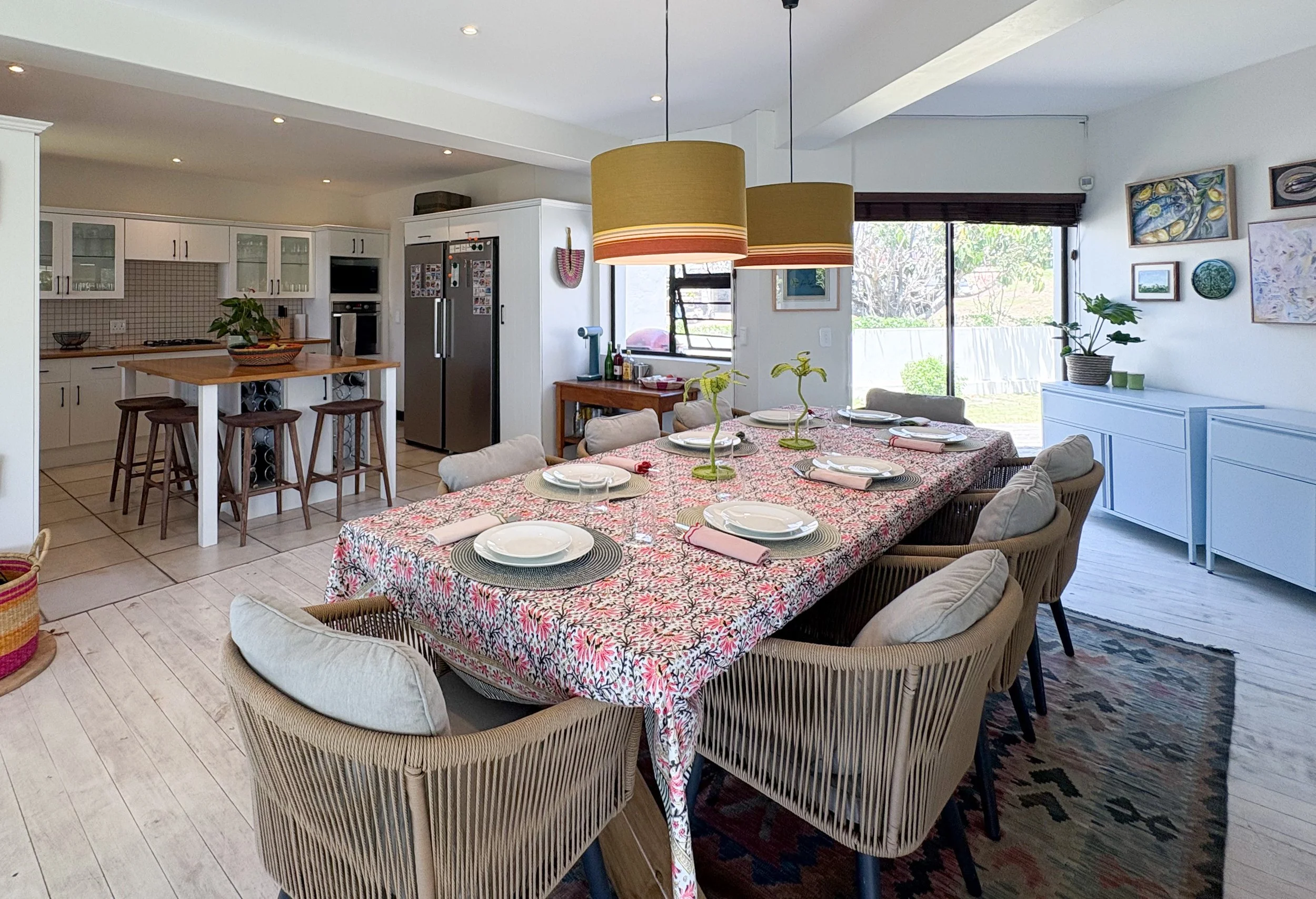 A dining room with a large table covered by a floral tablecloth, surrounded by wicker chairs with cushions. Overhead hang two large yellow pendant lights. The room features artwork on the white walls, a sideboard with plants, and large sliding glass doors letting in natural light, with a view of greenery outside.