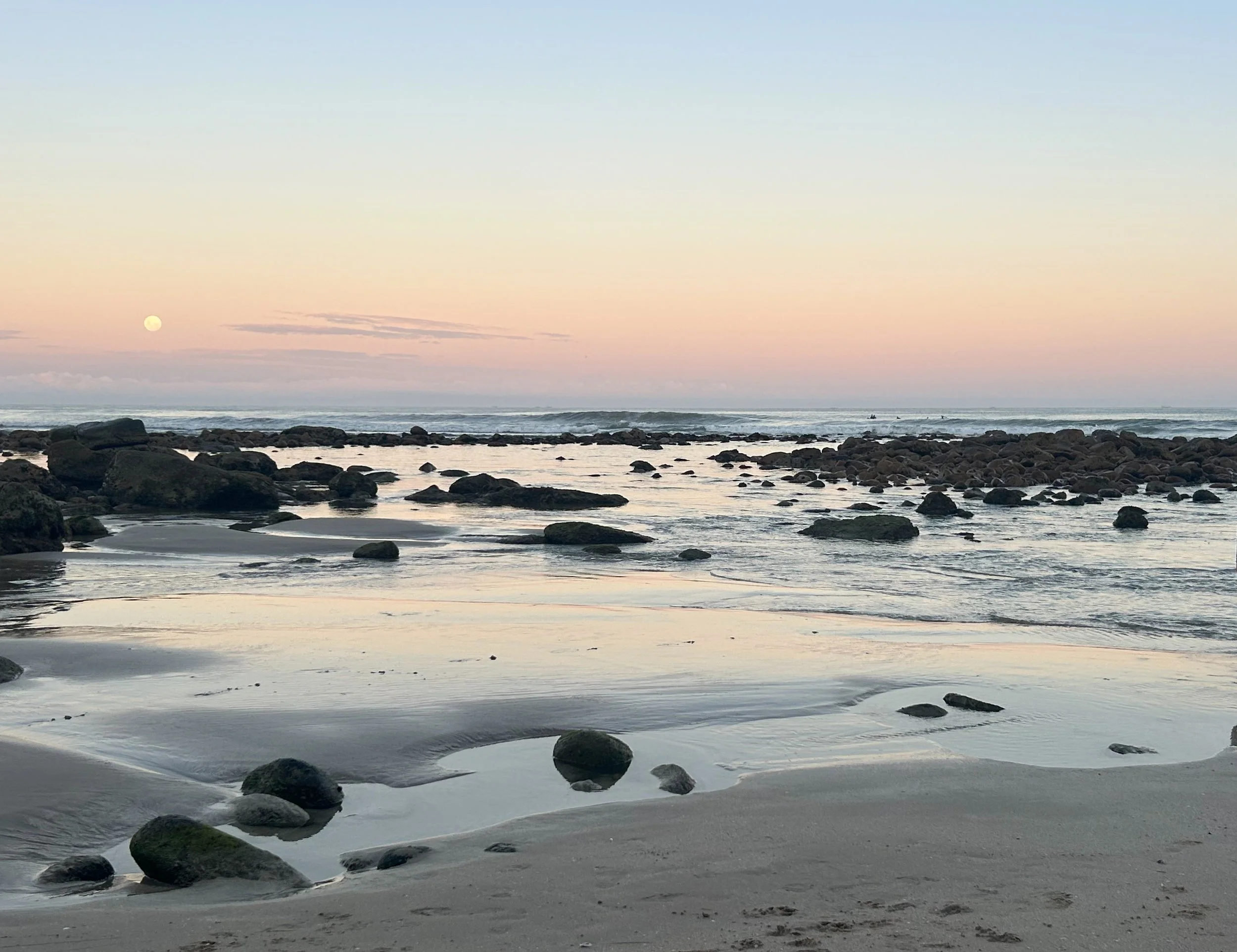 A serene beach scene during sunset with rocks scattered along the shoreline, gentle waves, and the moon visible in the sky.