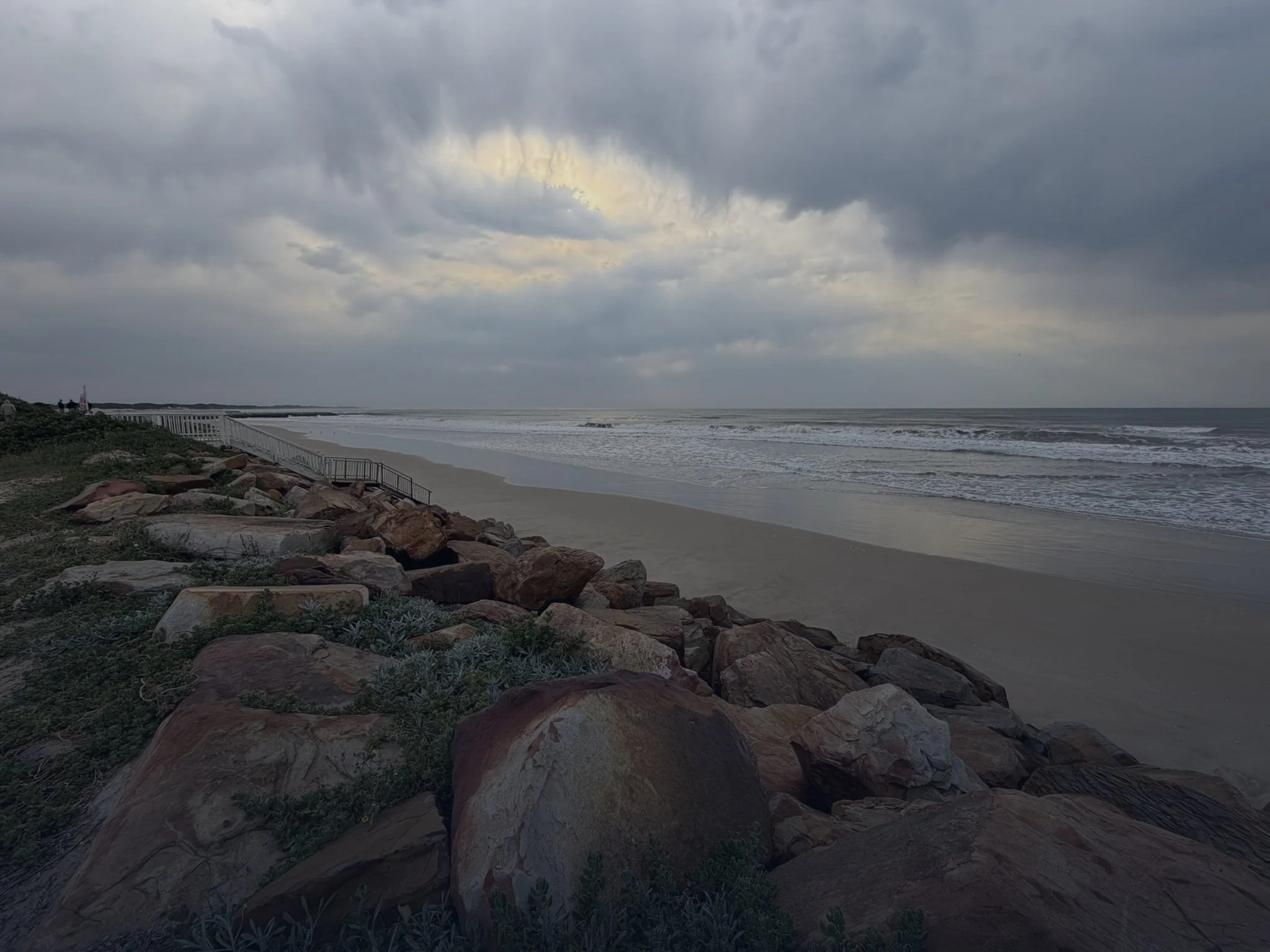 A view of a beach with sand, ocean waves, and a cloudy sky. Rocks and vegetation line the edge of the beach, with a small staircase leading down to the sand on the left side of the image.