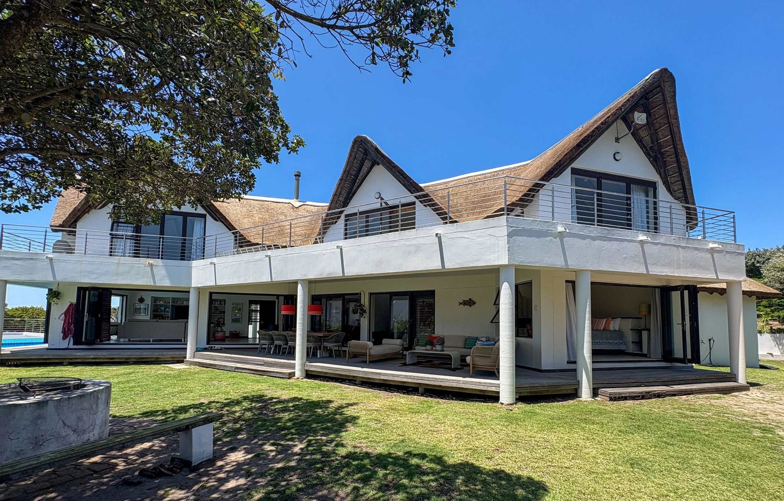 A modern house with a thatched roof, large glass sliding doors, and an upstairs balcony with metal railings. Outdoor seating is visible on the ground level, and a tree partially shades the yard with a grassy lawn.