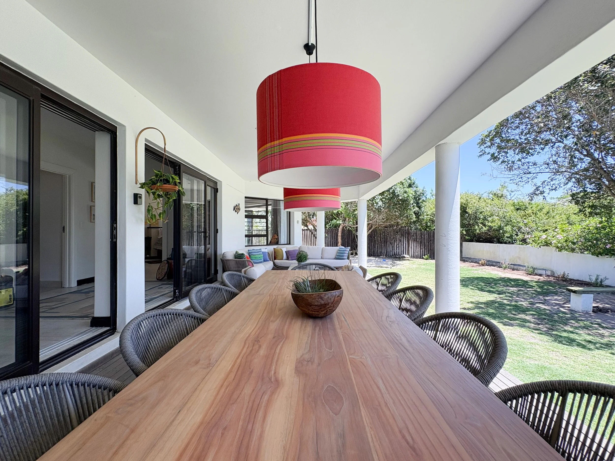 An outdoor patio with a wooden dining table surrounded by wicker chairs, a red hanging lantern, and a view of a backyard with trees and a white fence.